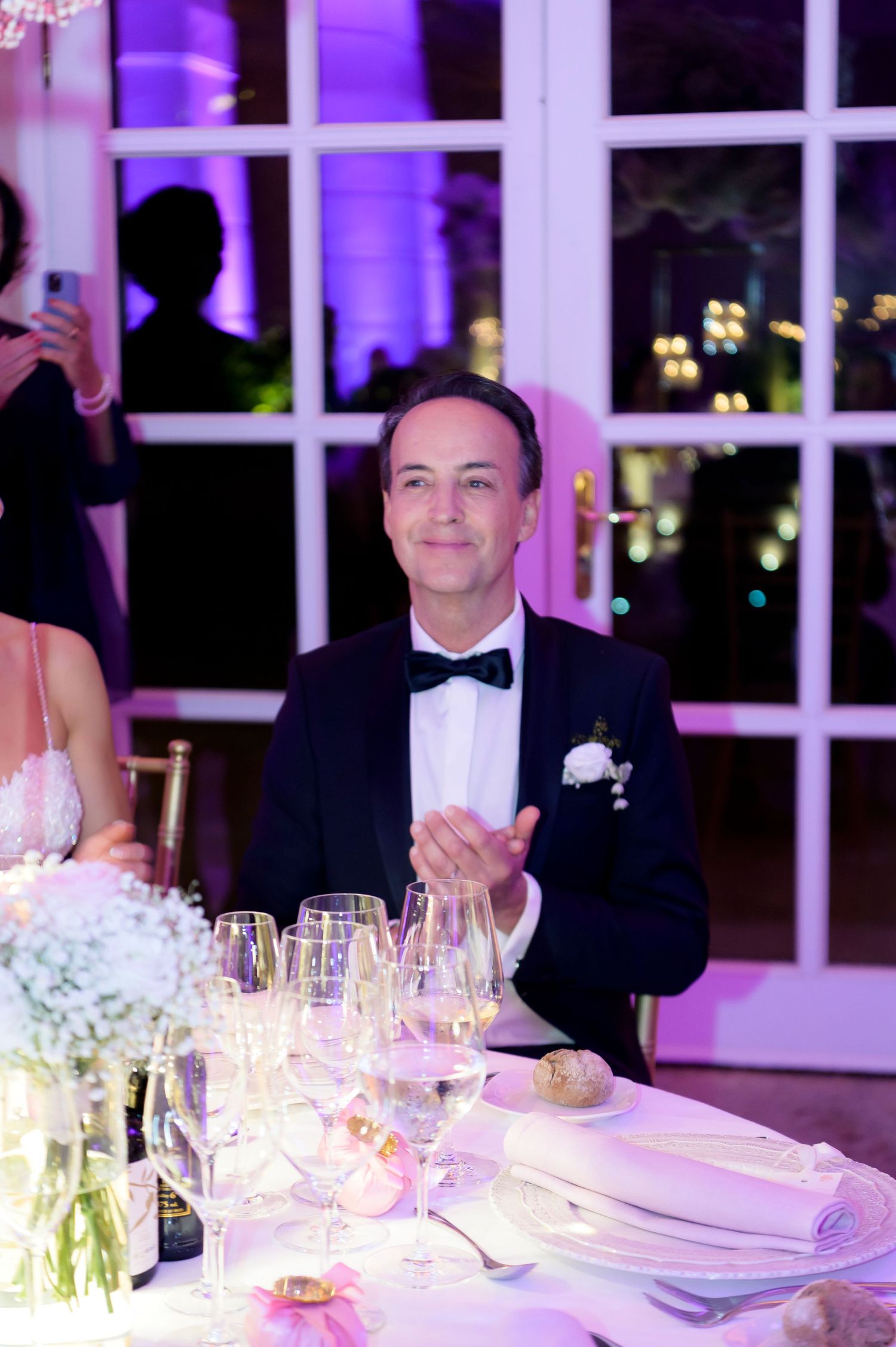 Smiling groom in a black tuxedo and bow tie sits at a wedding reception table with champagne glasses and floral centerpieces.