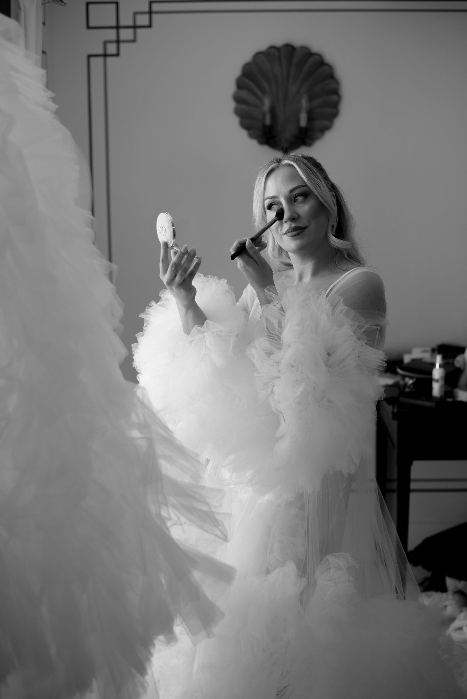 Black and white photo of a bride in a ruffled tulle robe applying makeup with a brush while holding a compact mirror in an elegant room.