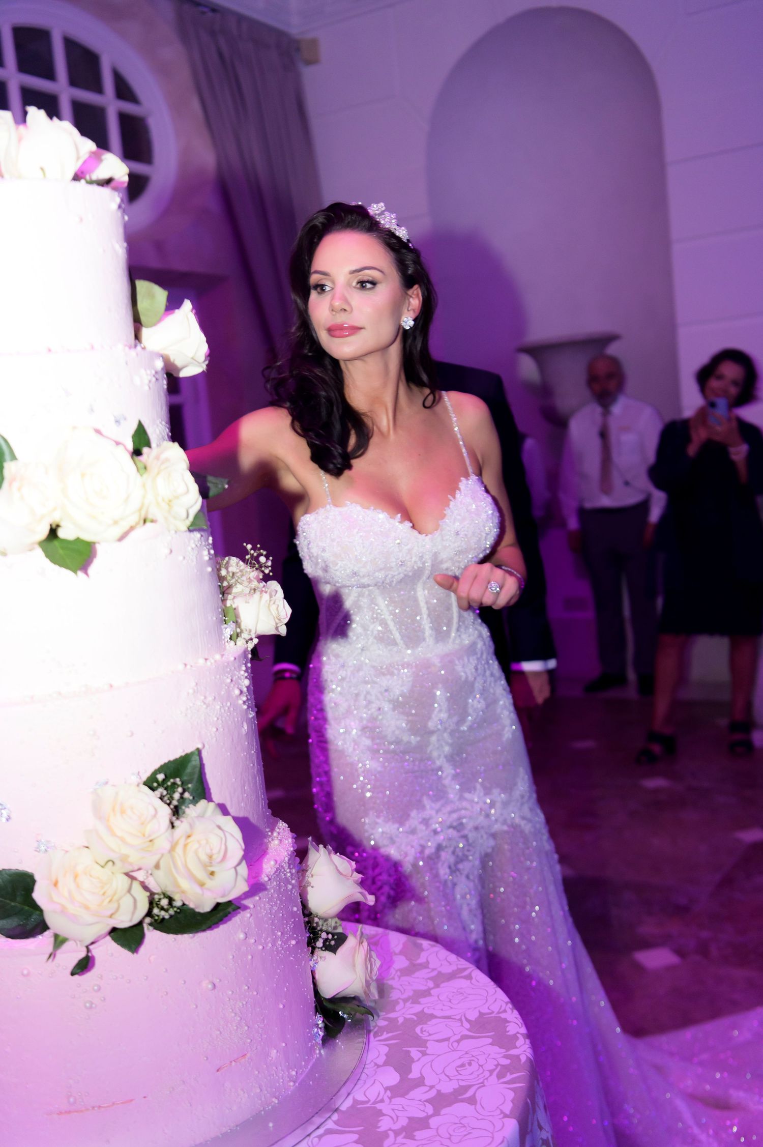 Bride in a sparkling lace wedding gown stands beside a towering white tiered wedding cake decorated with fresh roses.