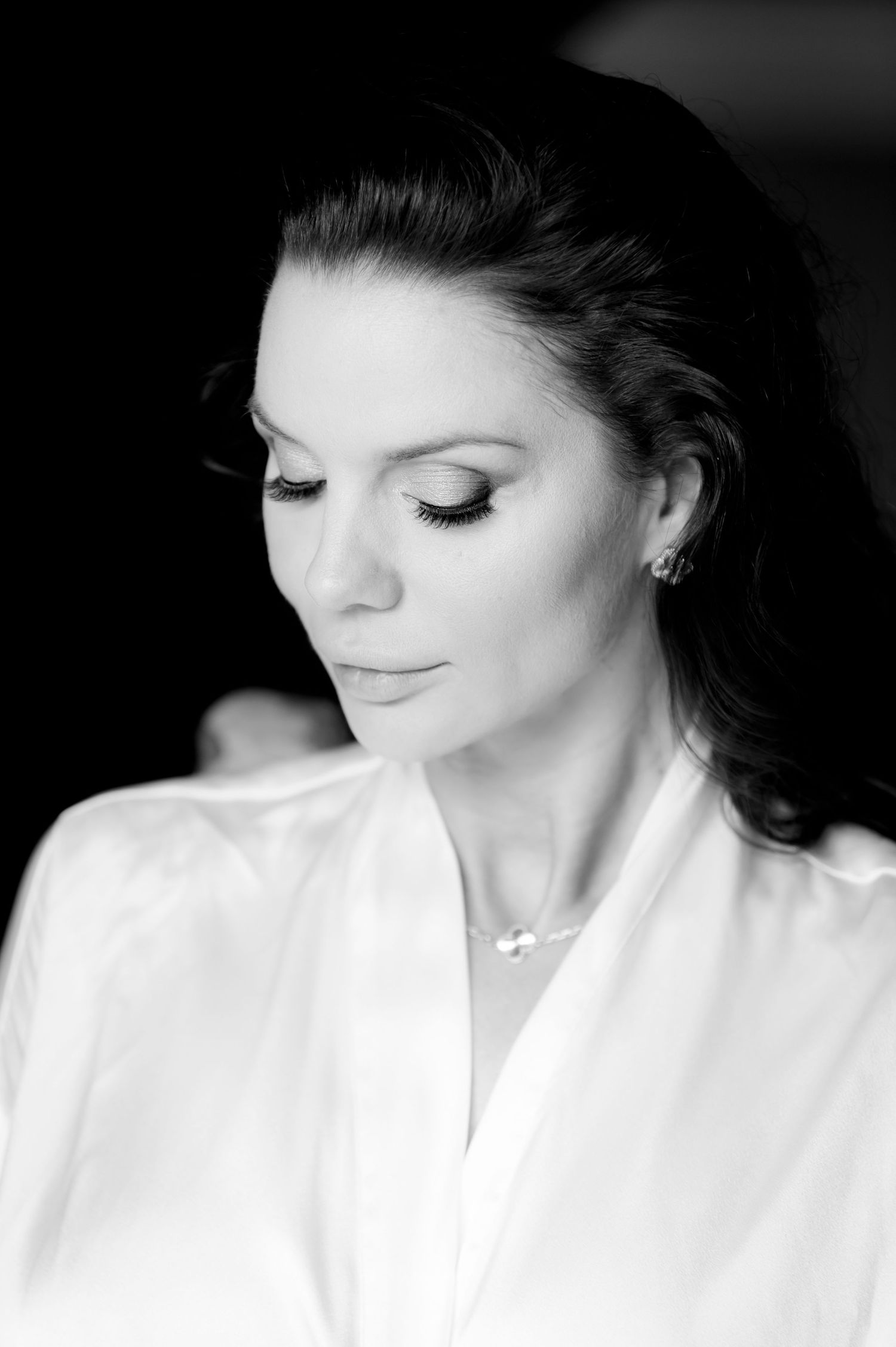 Black and white portrait of a woman in a white satin robe looking downward, with dark hair pulled back and soft makeup.