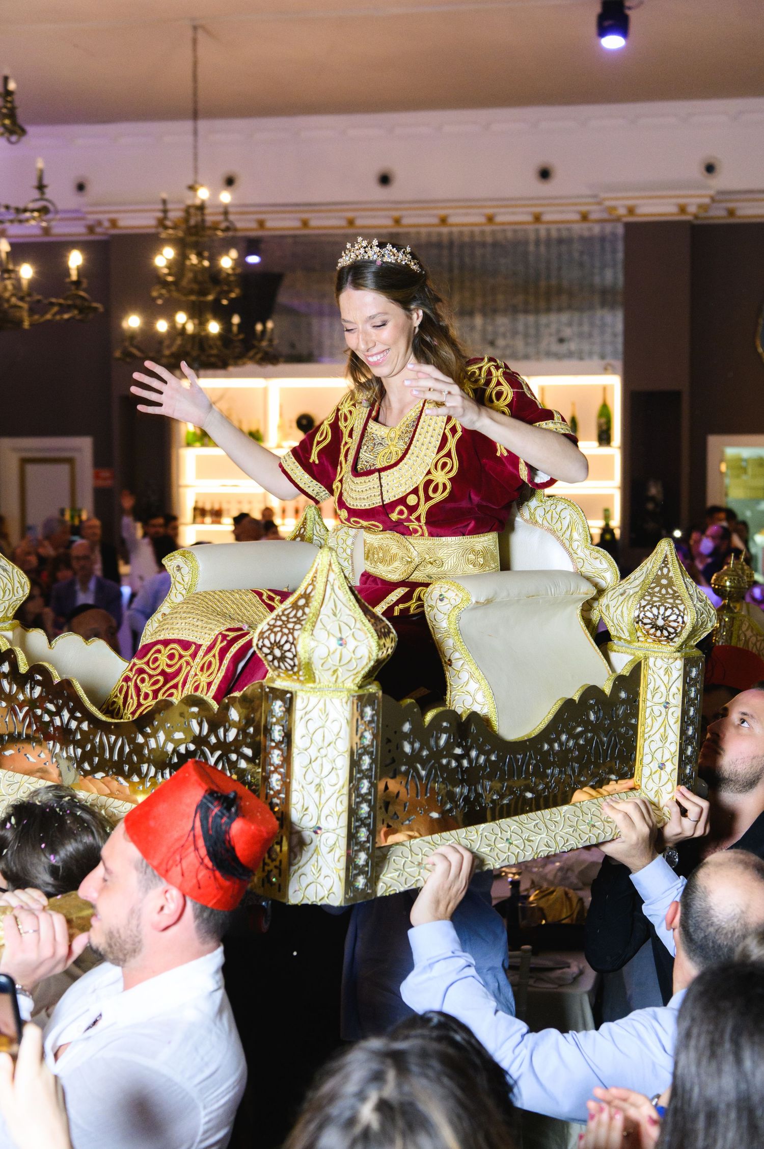 A smiling bride in a red and gold embroidered dress and tiara is lifted on an ornate decorated chair by cheering wedding guests.