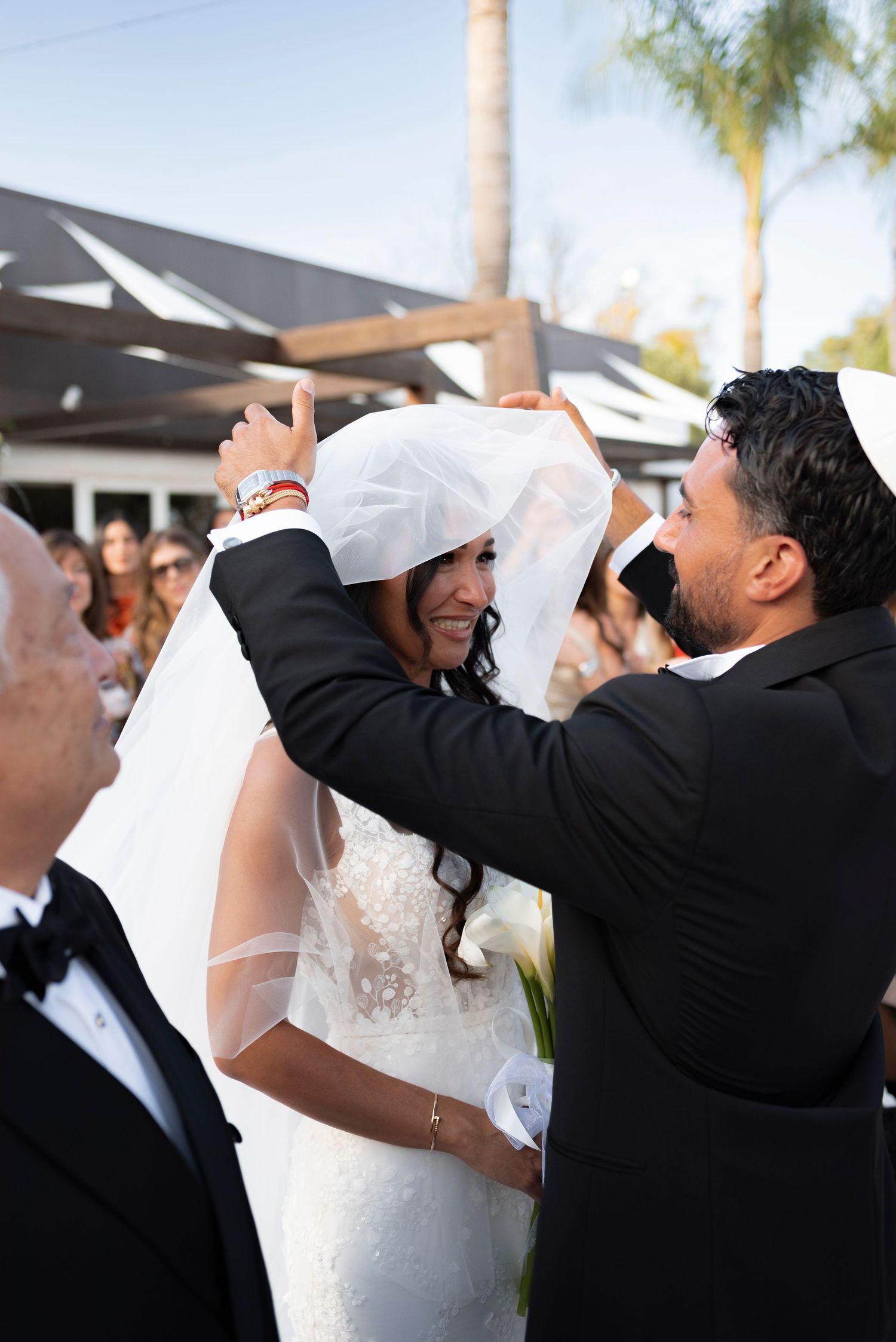 Groom in a kippah lifts the veil over a smiling bride in a lace gown during an outdoor wedding ceremony as guests look on.
