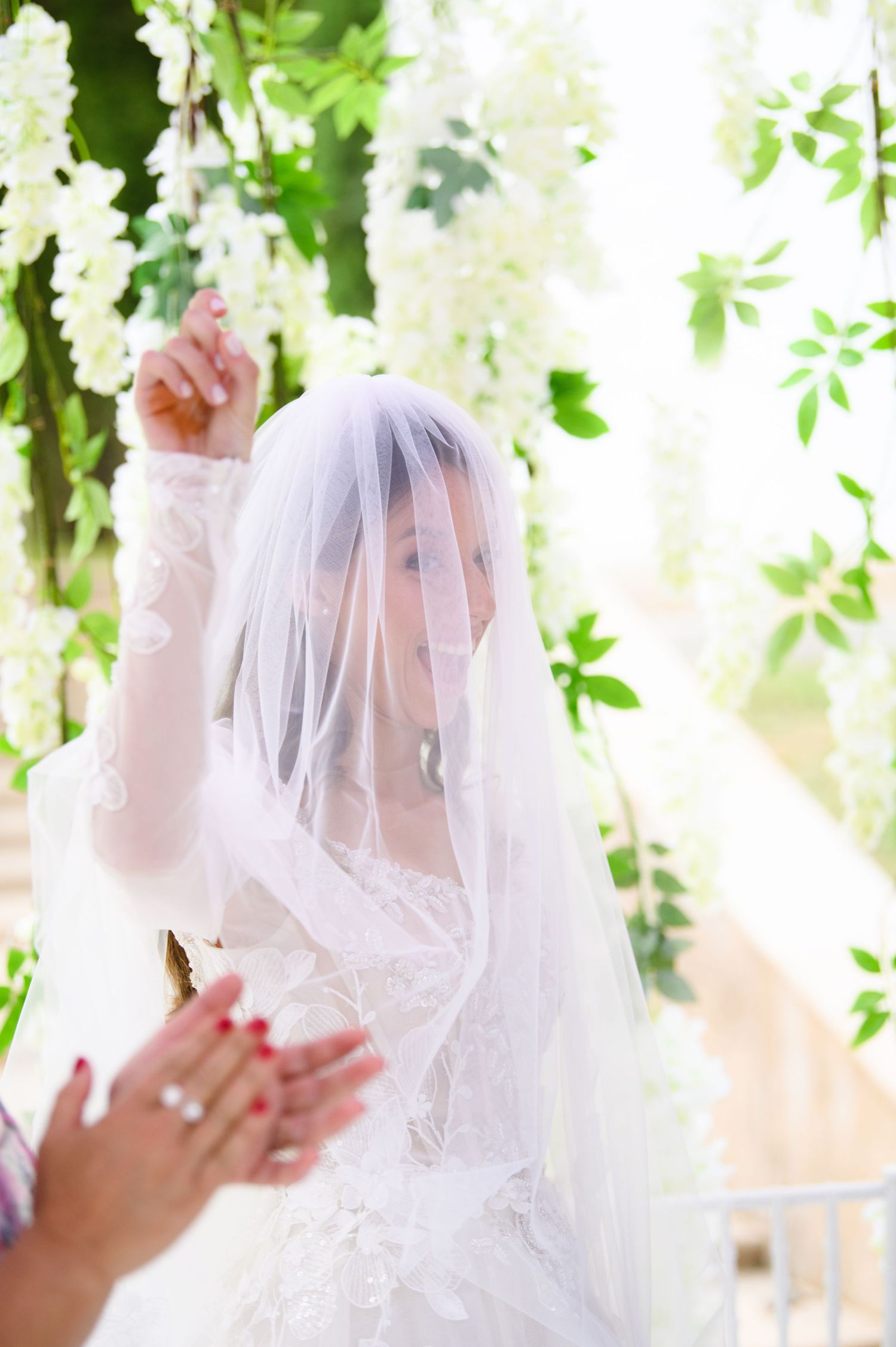 Bride in a lace wedding gown and sheer veil raises her hand beneath hanging white wisteria blossoms at an outdoor ceremony.