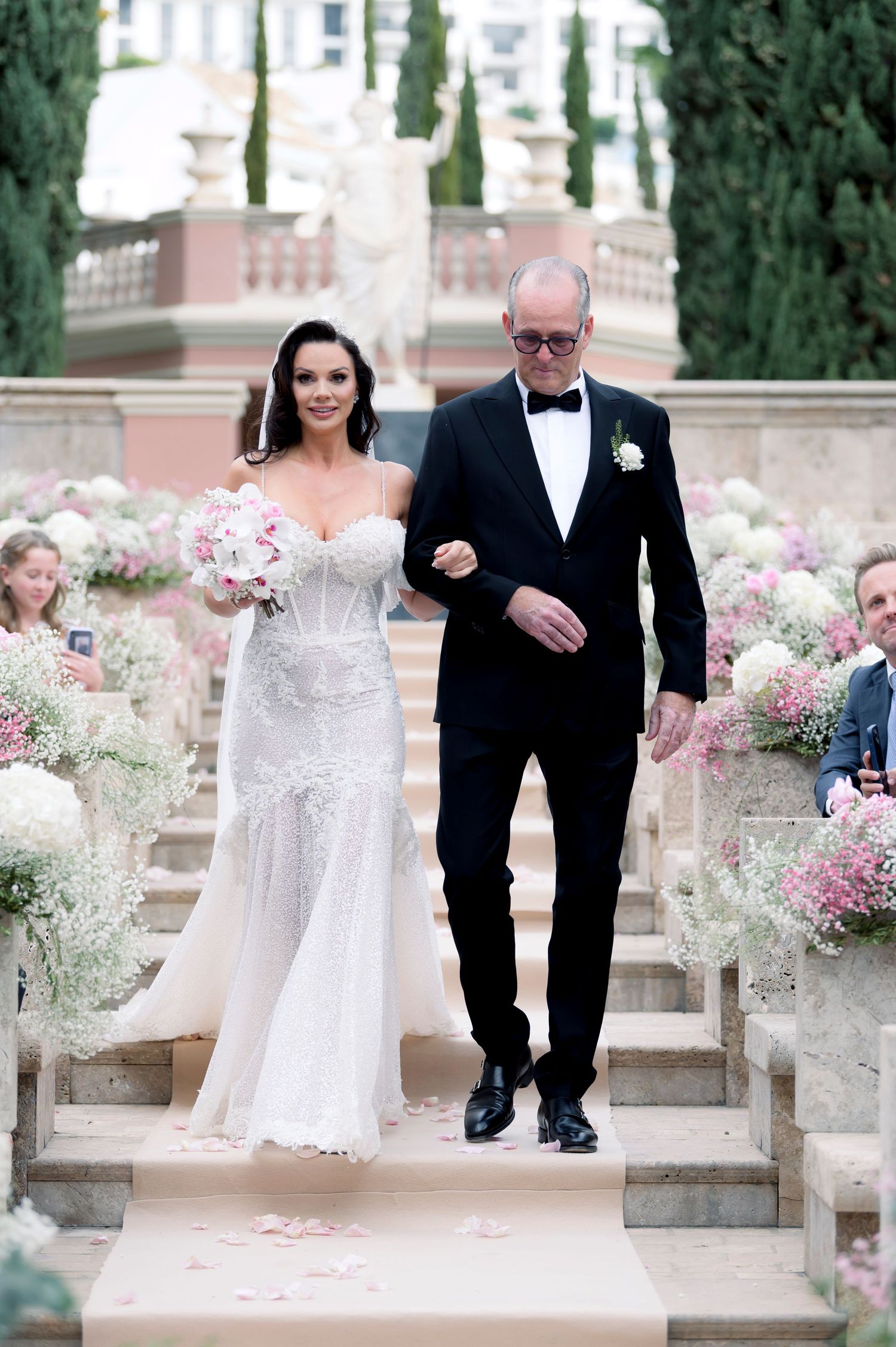 A bride in a lace mermaid wedding gown carries a bouquet while walking down garden stairs on the arm of a man in a black tuxedo.