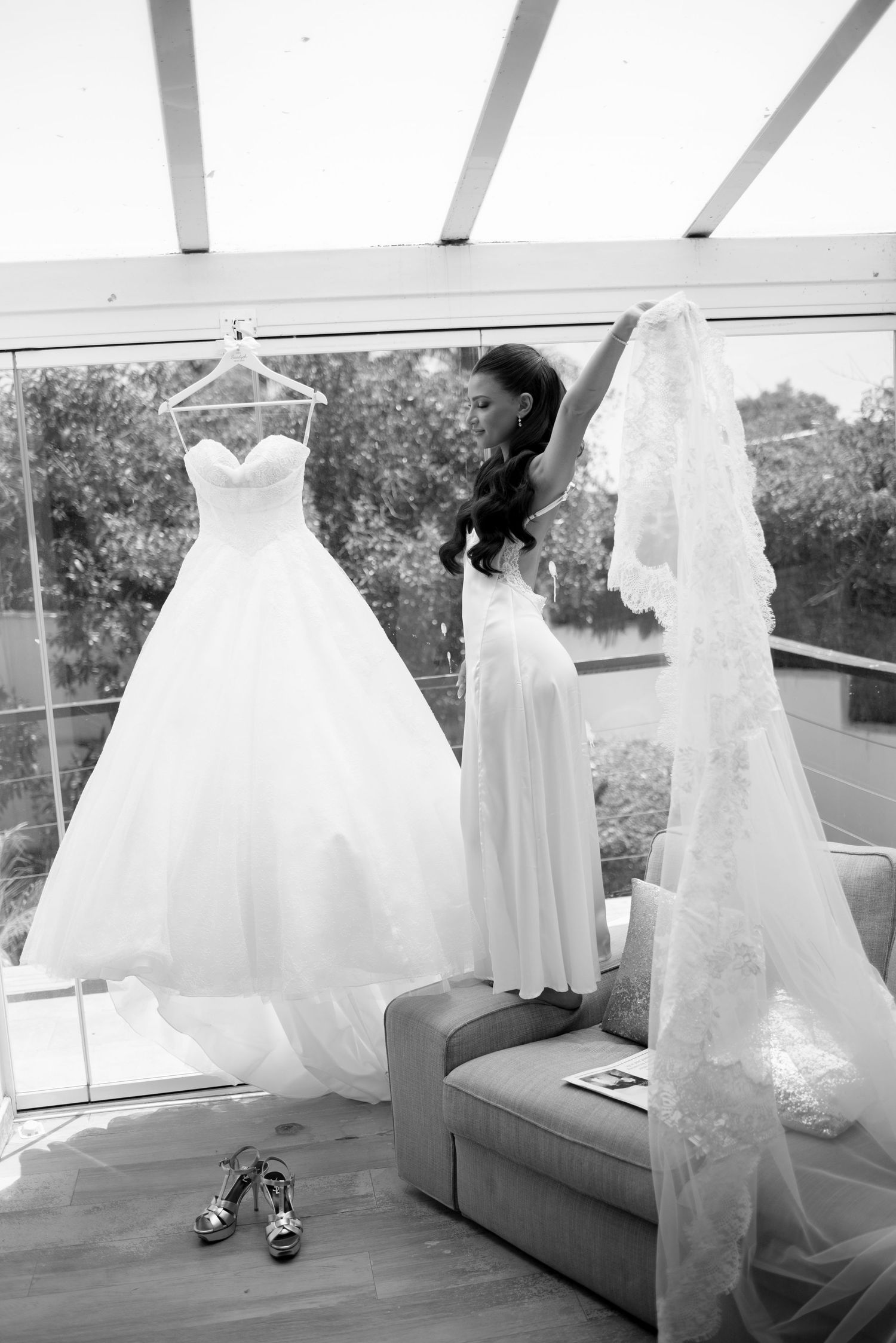 Bride in a slip stands on a sofa, lifting a long lace veil beside her wedding gown hanging by a window, with heels on the floor.