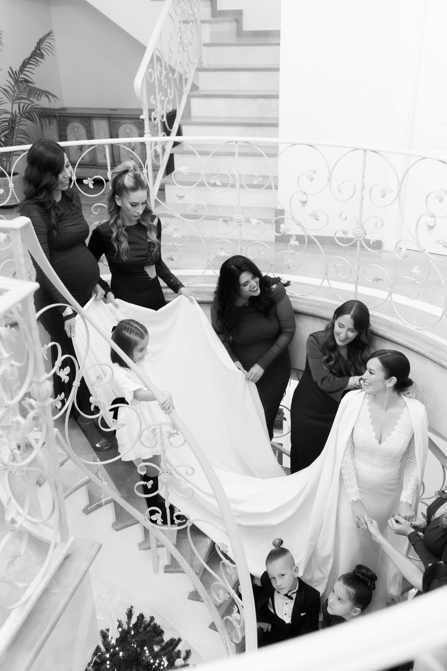 Black and white photo of a bride in a long gown on a curved staircase as bridesmaids and children hold her flowing veil.