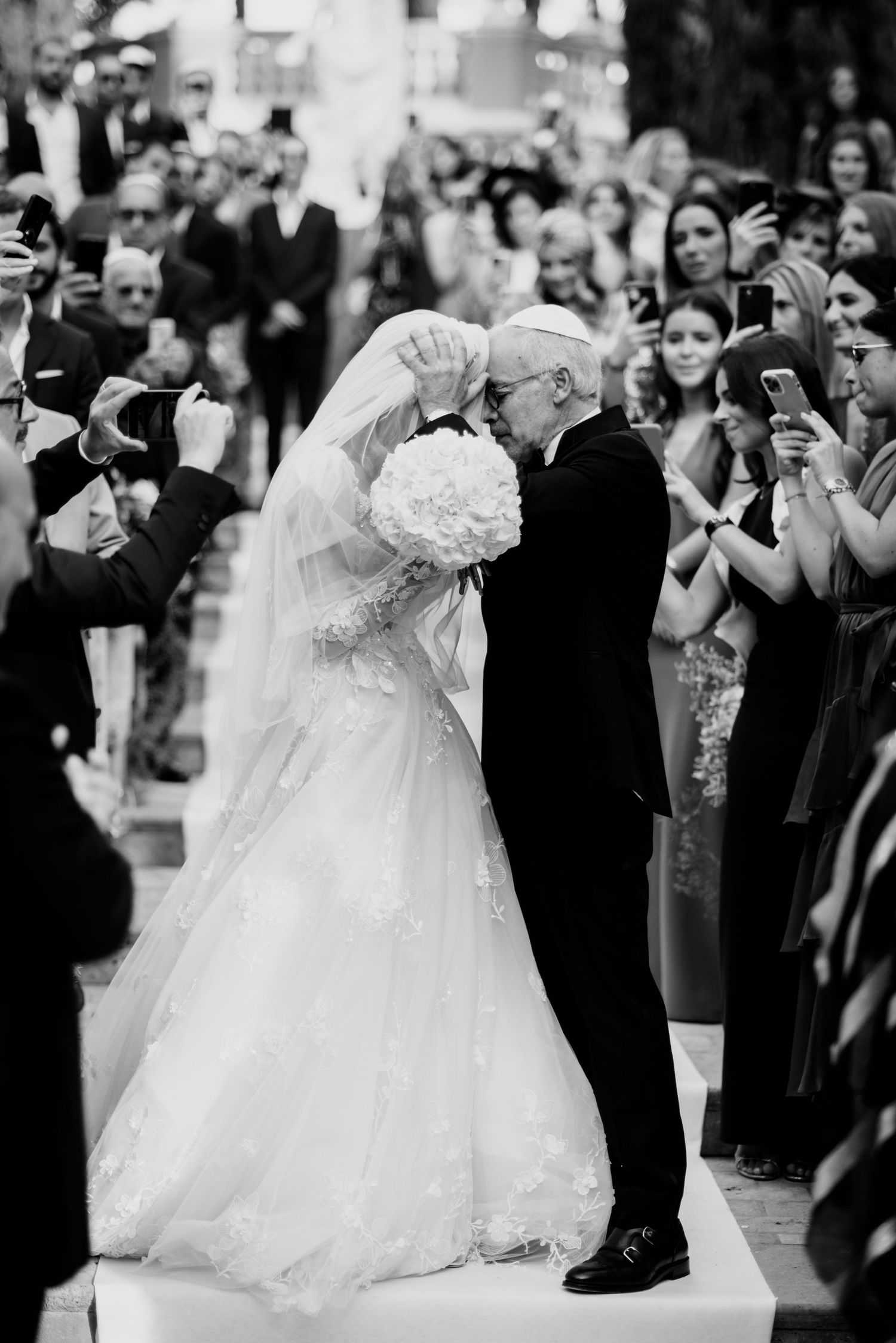 A bride in a lace gown and long veil holds a white bouquet as an older man in a dark suit embraces her during a wedding ceremony.