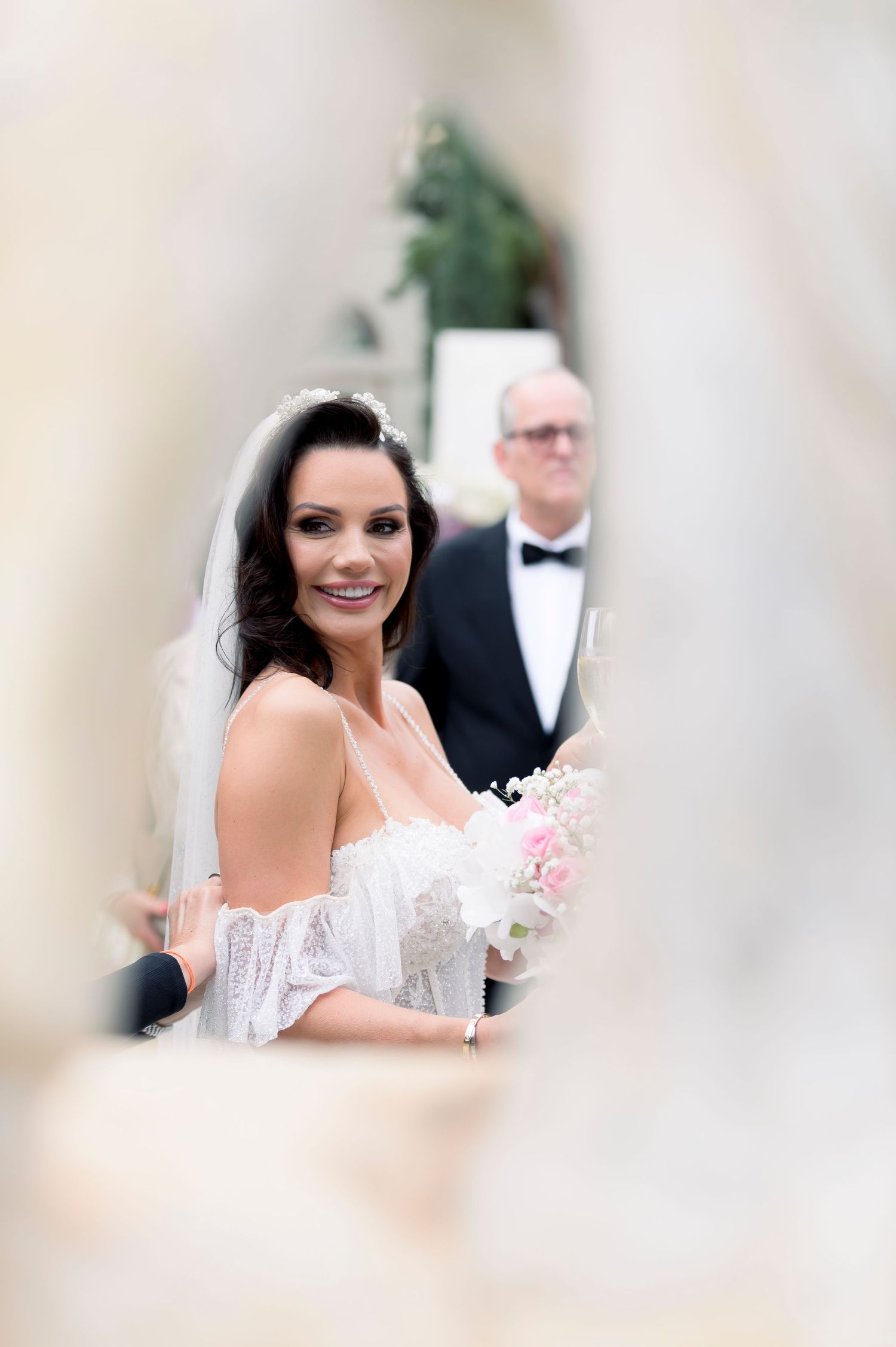 Smiling bride in an off-shoulder lace wedding gown and veil holds a pink bouquet as a man in a tuxedo and bow tie stands behind her.