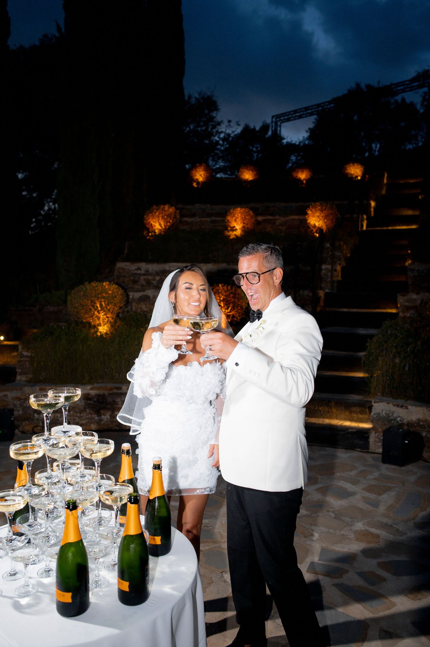 Bride in a short white dress and veil toasts with a groom in a white dinner jacket beside a champagne tower at an evening garden wedding.