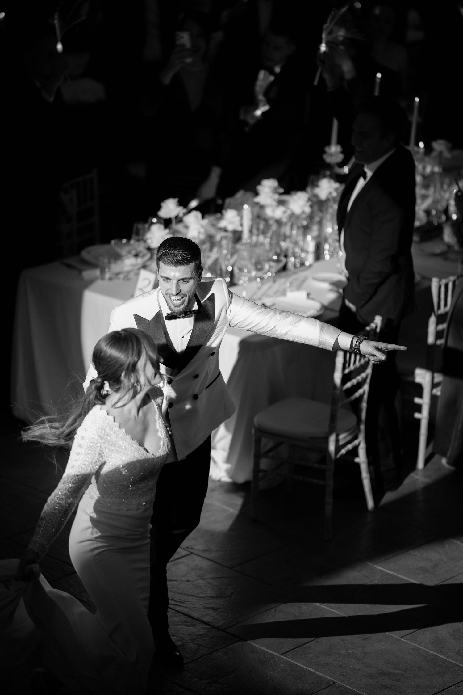 Smiling groom in a vest and bow tie dances with his bride in a flowing gown beside elegantly set reception tables in black and white.