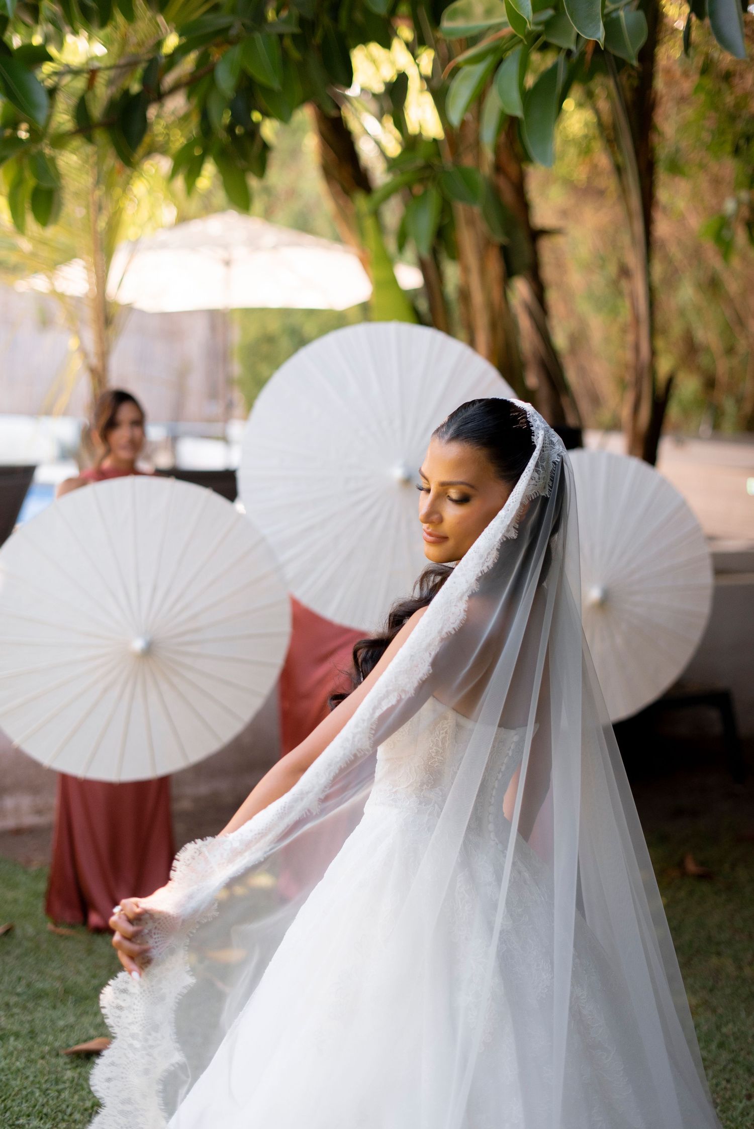 Bride in a white lace-trimmed wedding gown and long veil holds the fabric outdoors beneath white paper parasols and green trees.