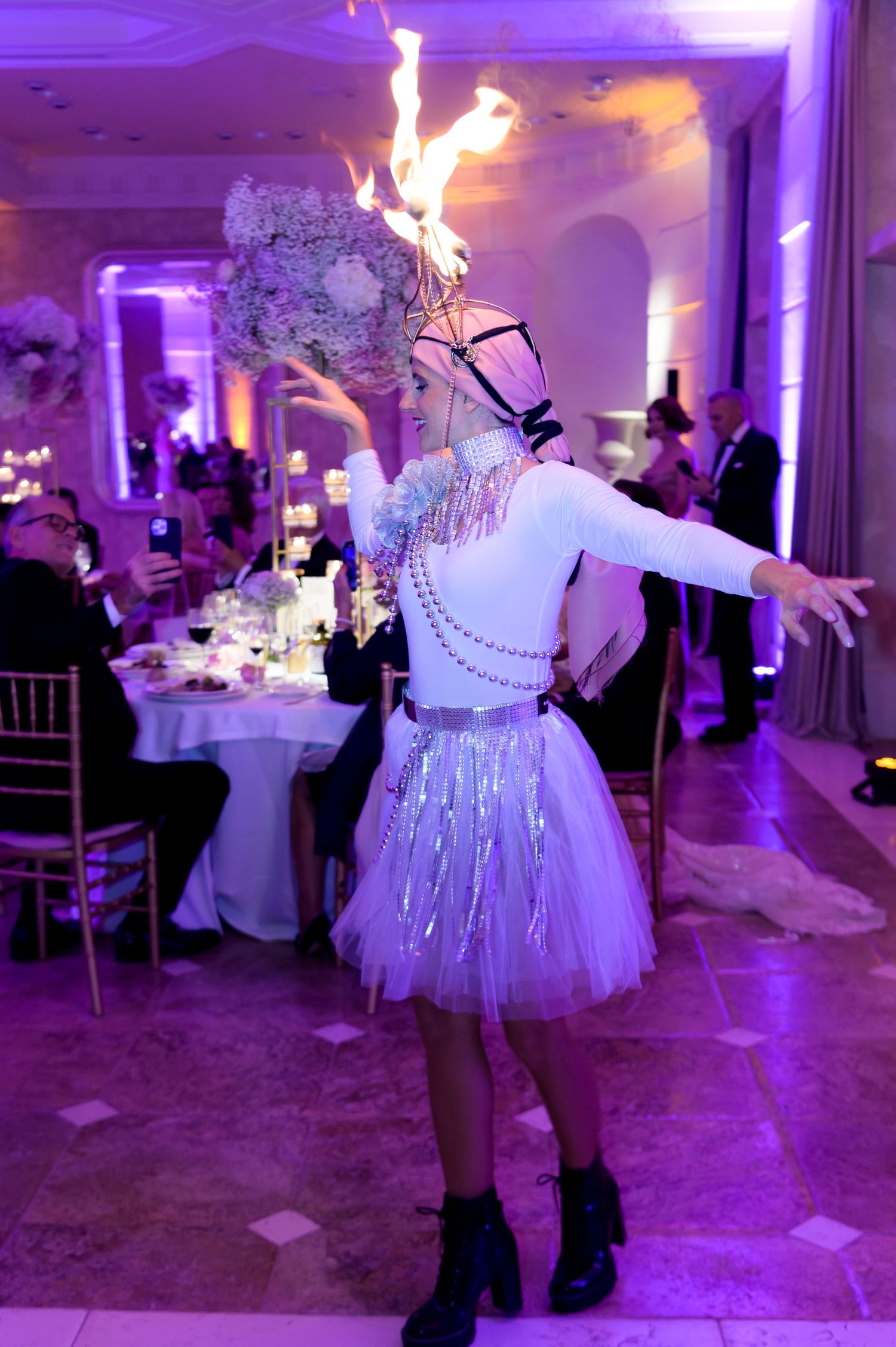 Performer in a flame headpiece, white top and sparkling skirt dances through an elegant banquet hall with guests seated at floral tables.