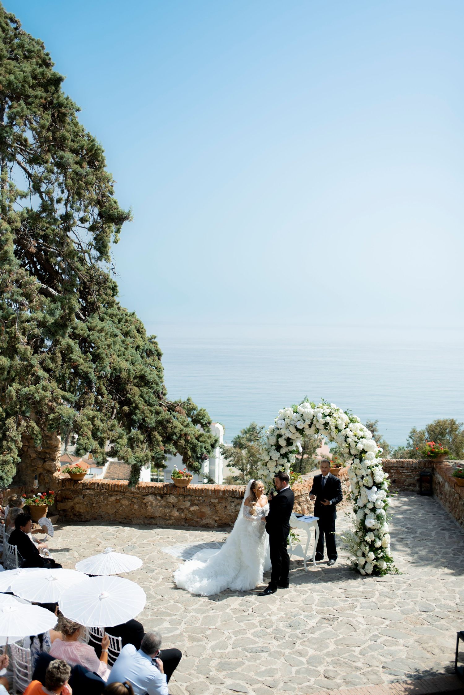 Bride and groom exchange vows beneath a white floral arch at a seaside stone terrace wedding as guests watch under umbrellas.
