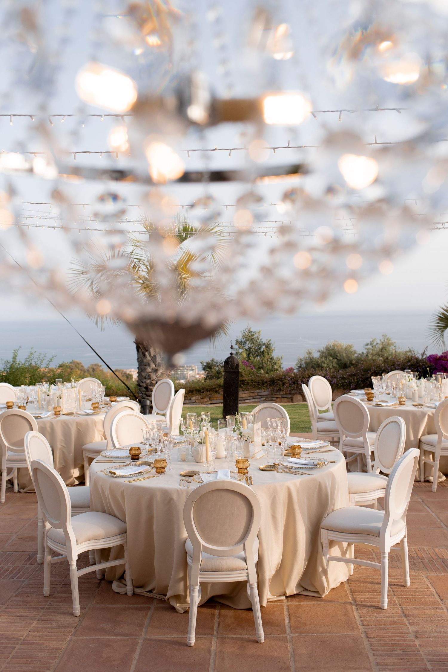 Elegant outdoor wedding reception with round tables, white chairs and a crystal chandelier overlooking the ocean at sunset.