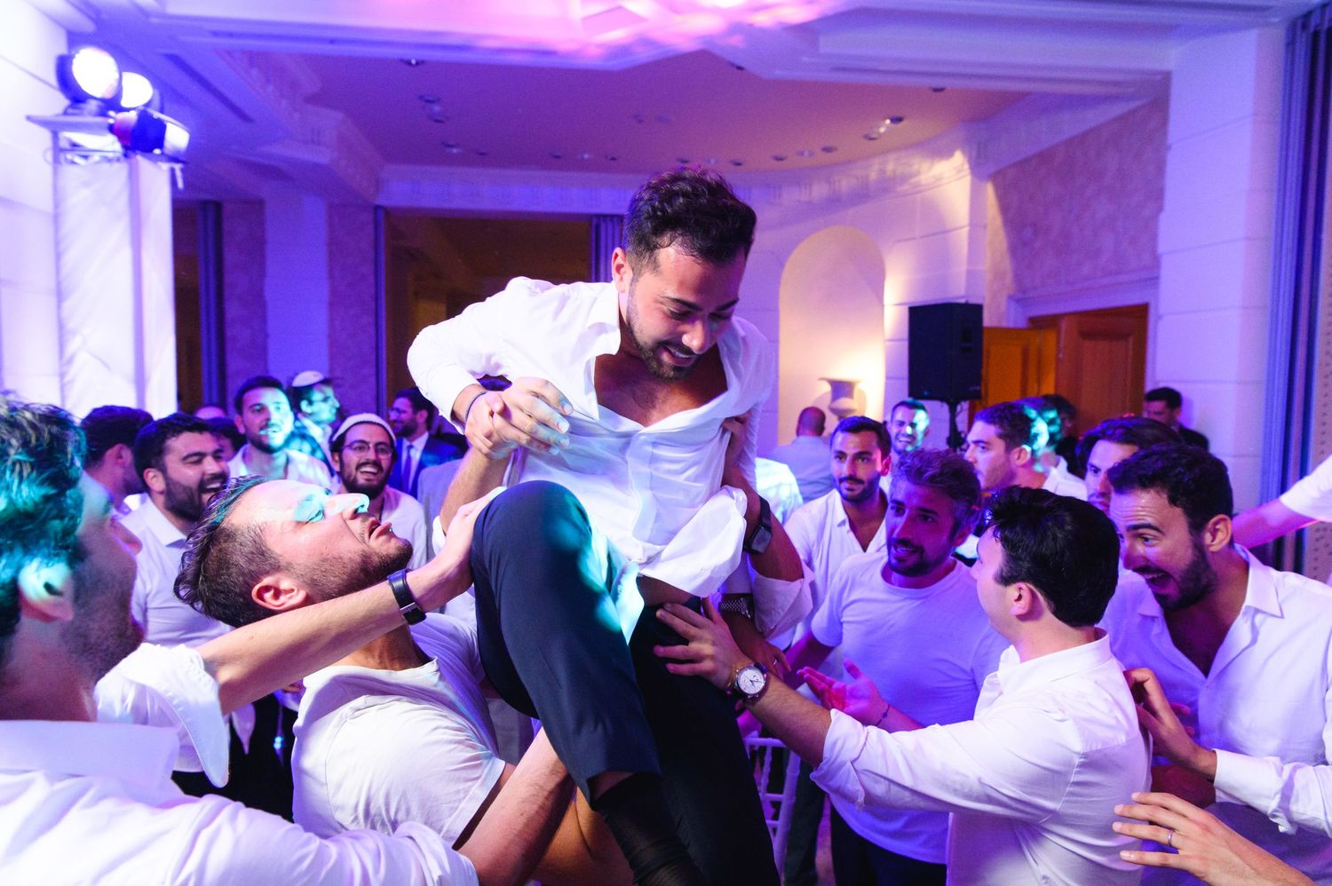 Cheerful wedding guests in white shirts lift a smiling man into the air on their shoulders while celebrating on a lively dance floor.