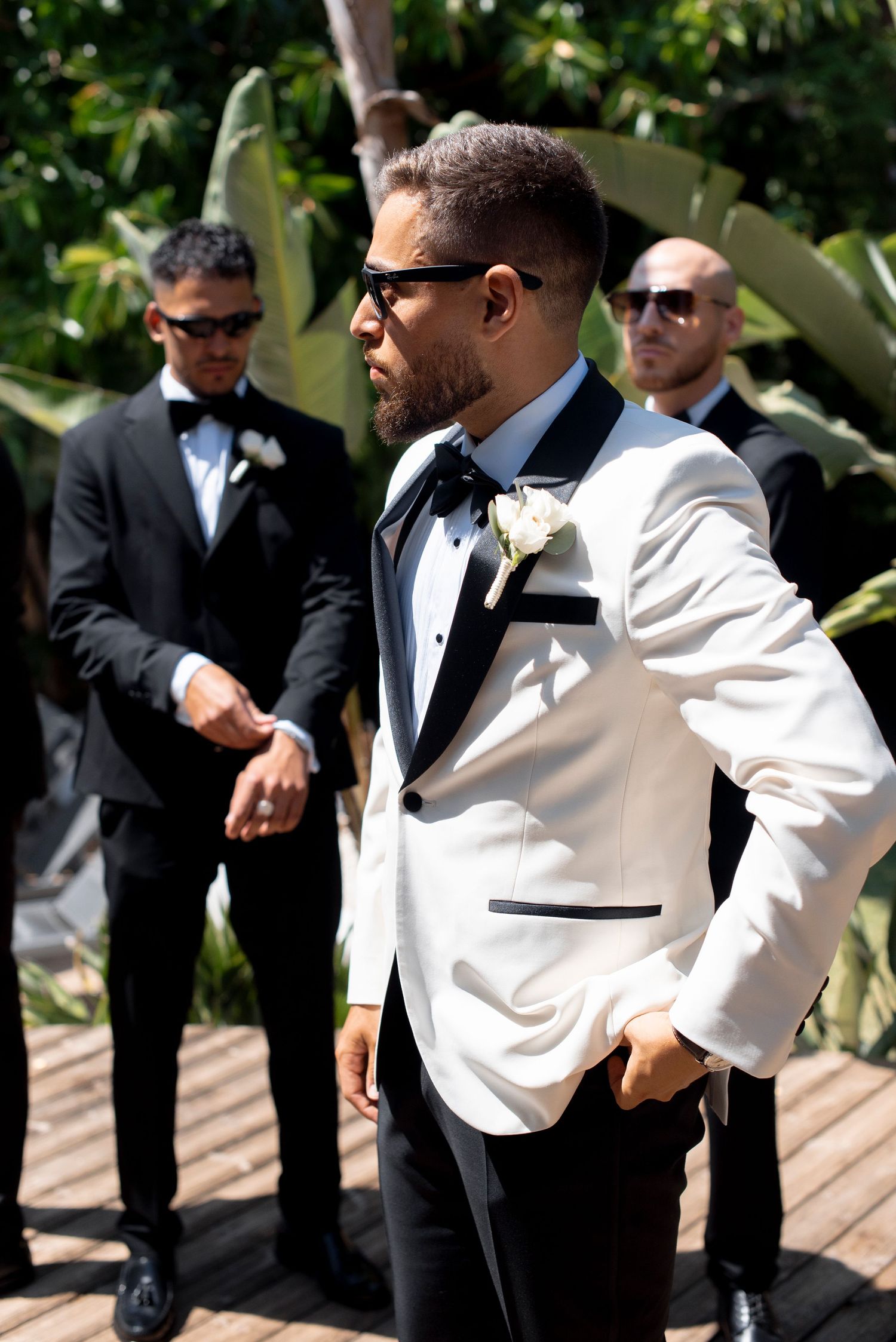 Groom in a white tuxedo jacket, black bow tie and sunglasses stands on a wooden deck with groomsmen in black suits behind him.