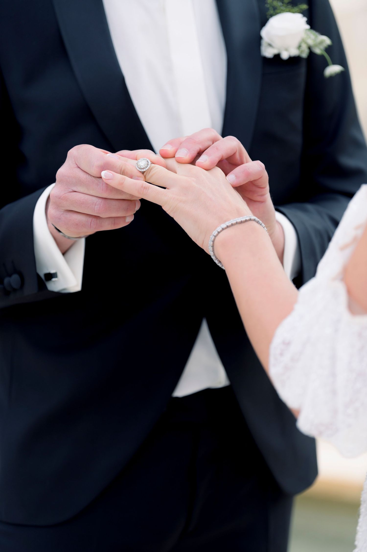 Groom in a black tuxedo with a white boutonniere slides a diamond wedding ring onto the bride's finger during the ceremony.