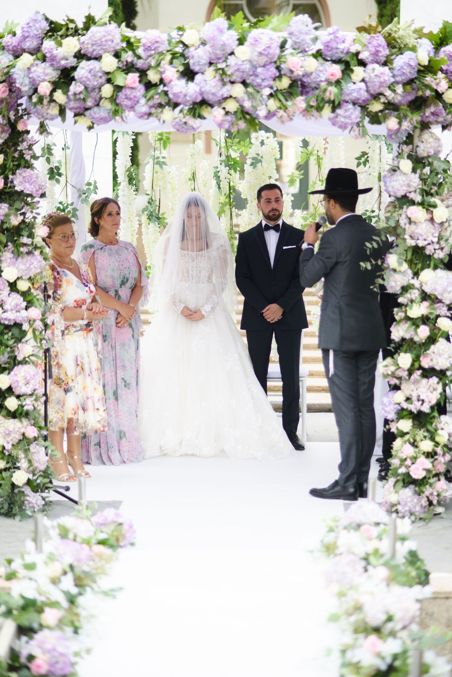 Bride in a lace gown and veil stands beside the groom under a lavender floral wedding canopy as an officiant leads the ceremony.