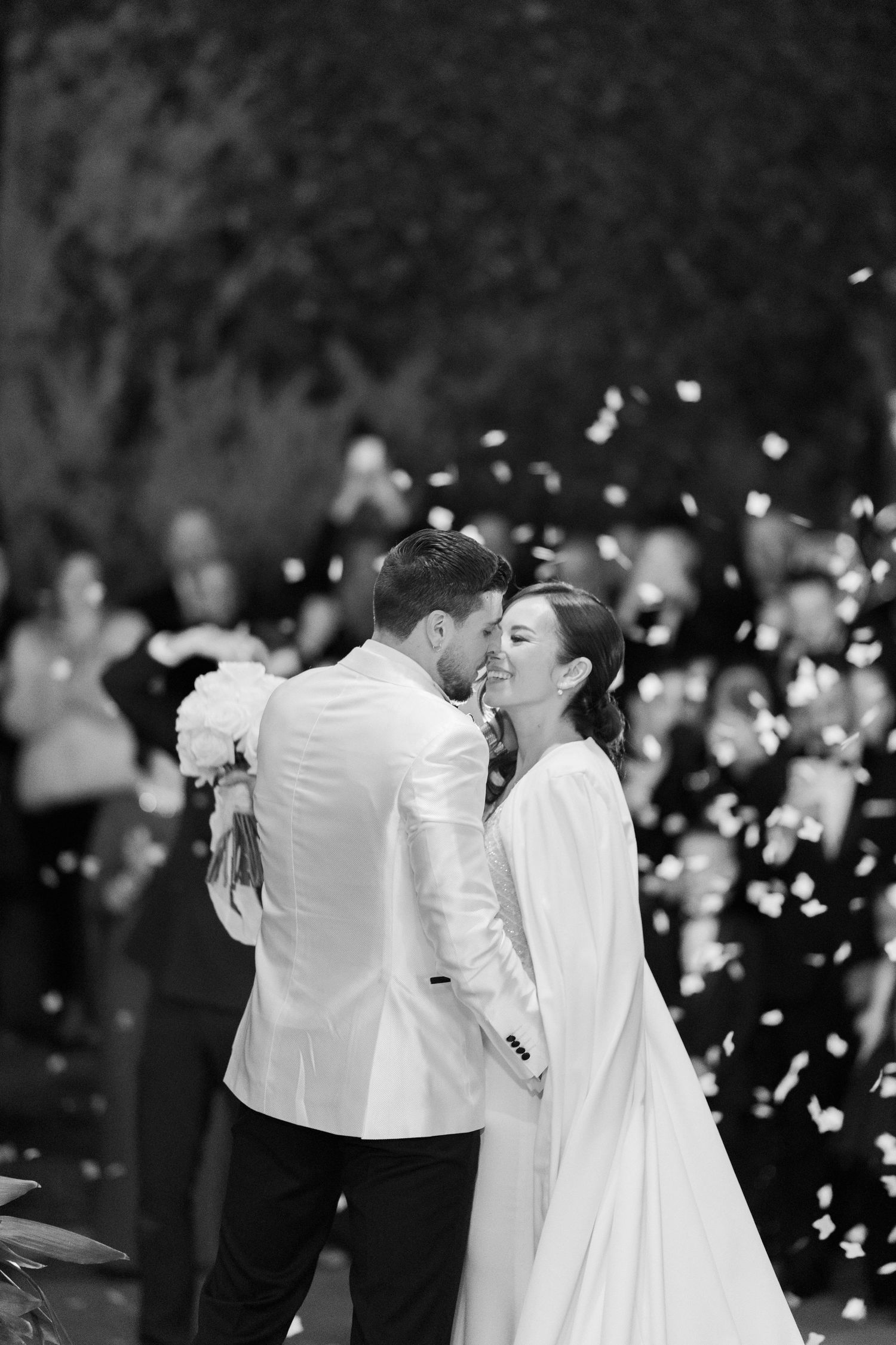 Black and white photo of a bride and groom kissing as guests toss confetti around them at their outdoor wedding ceremony.