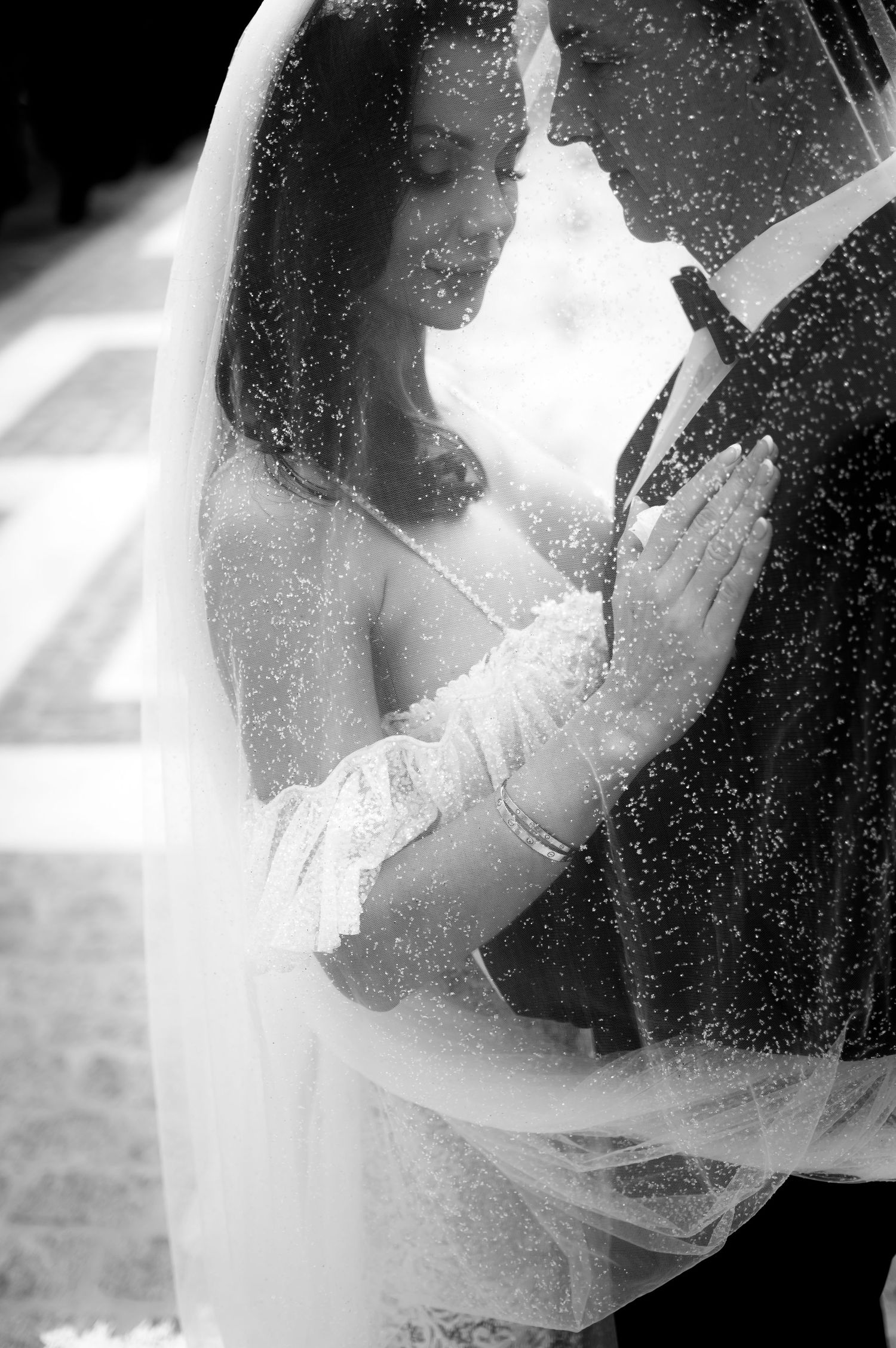 Black and white photo of a bride and groom embracing under a sheer veil, her hand resting on his suit lapel.
