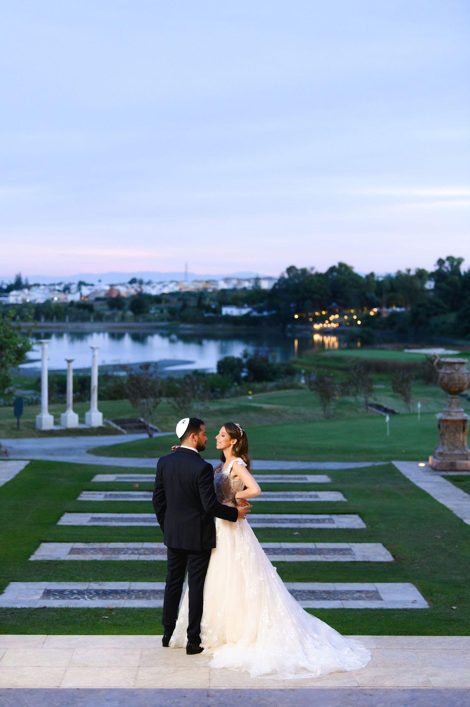 A bride in a flowing white gown and a groom in a dark suit and kippah embrace on stone steps overlooking a lake at dusk.