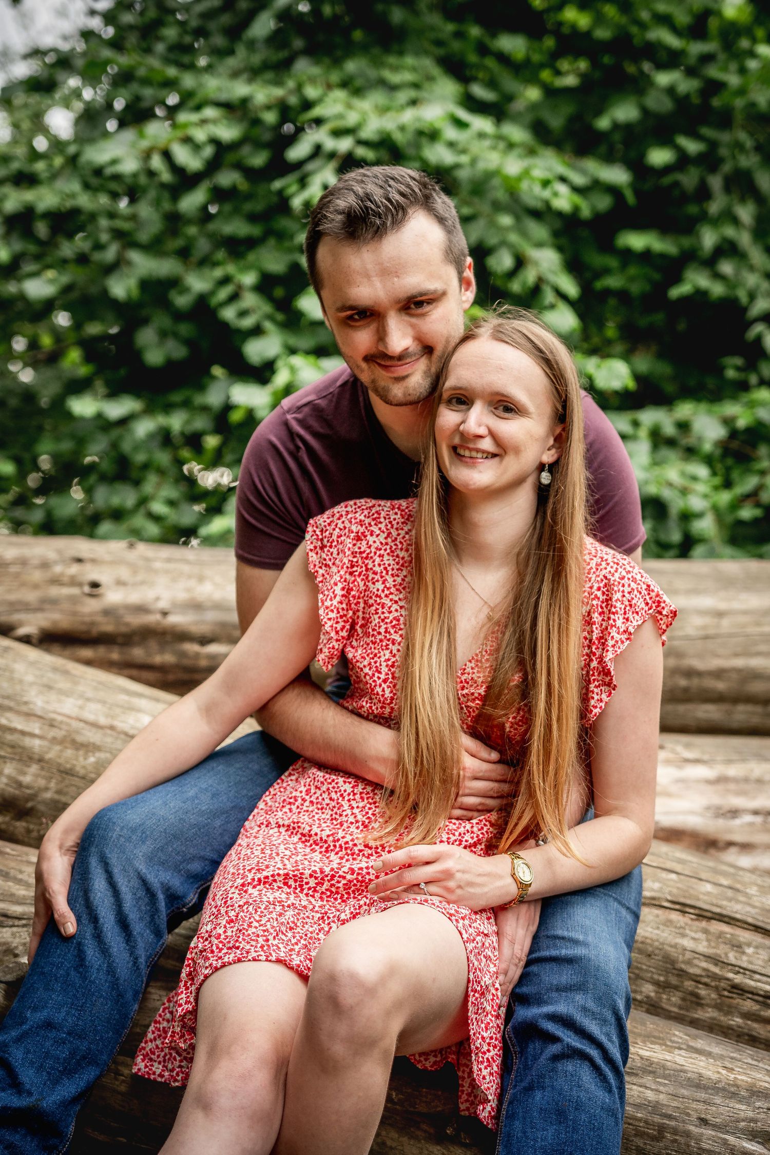 couple sitting on a pile of logs and embracing, woman sitting between mans' legs, looking at us