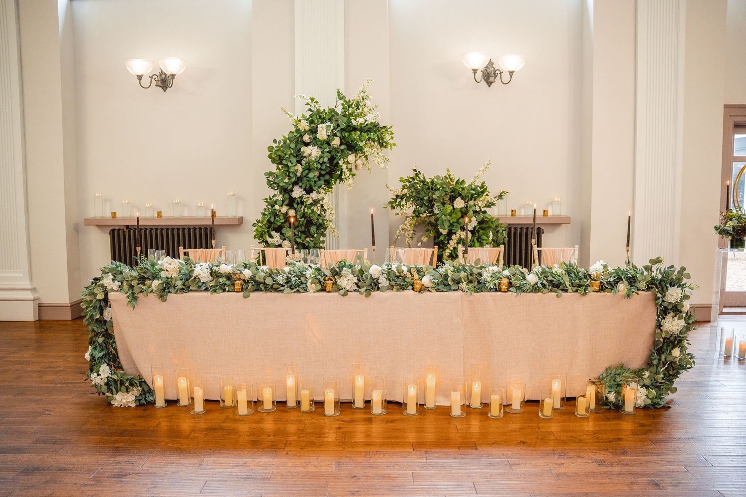 Top table with pink table cloth, greenery and white flowers garland laid on table, candles lined along the base of table