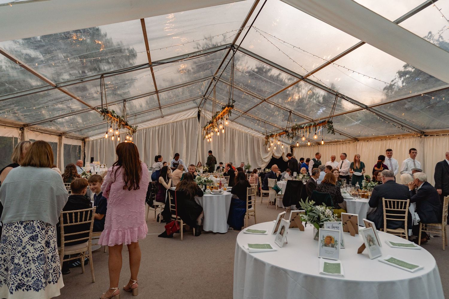 Elegant outdoor wedding reception under a clear tent with string lights, round tables, and guests mingling at dusk.