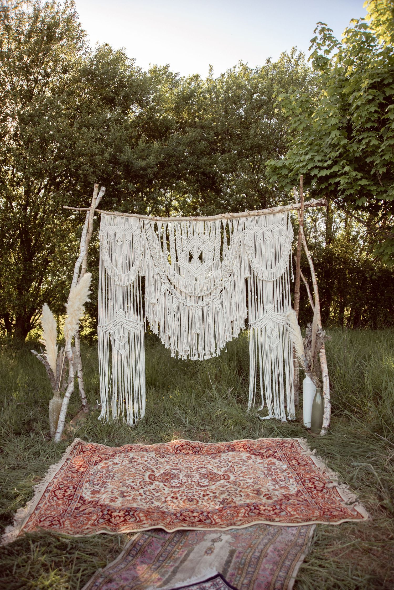 Macrame wedding backdrop hanging over a vintage patterned rug in an outdoor garden setting.