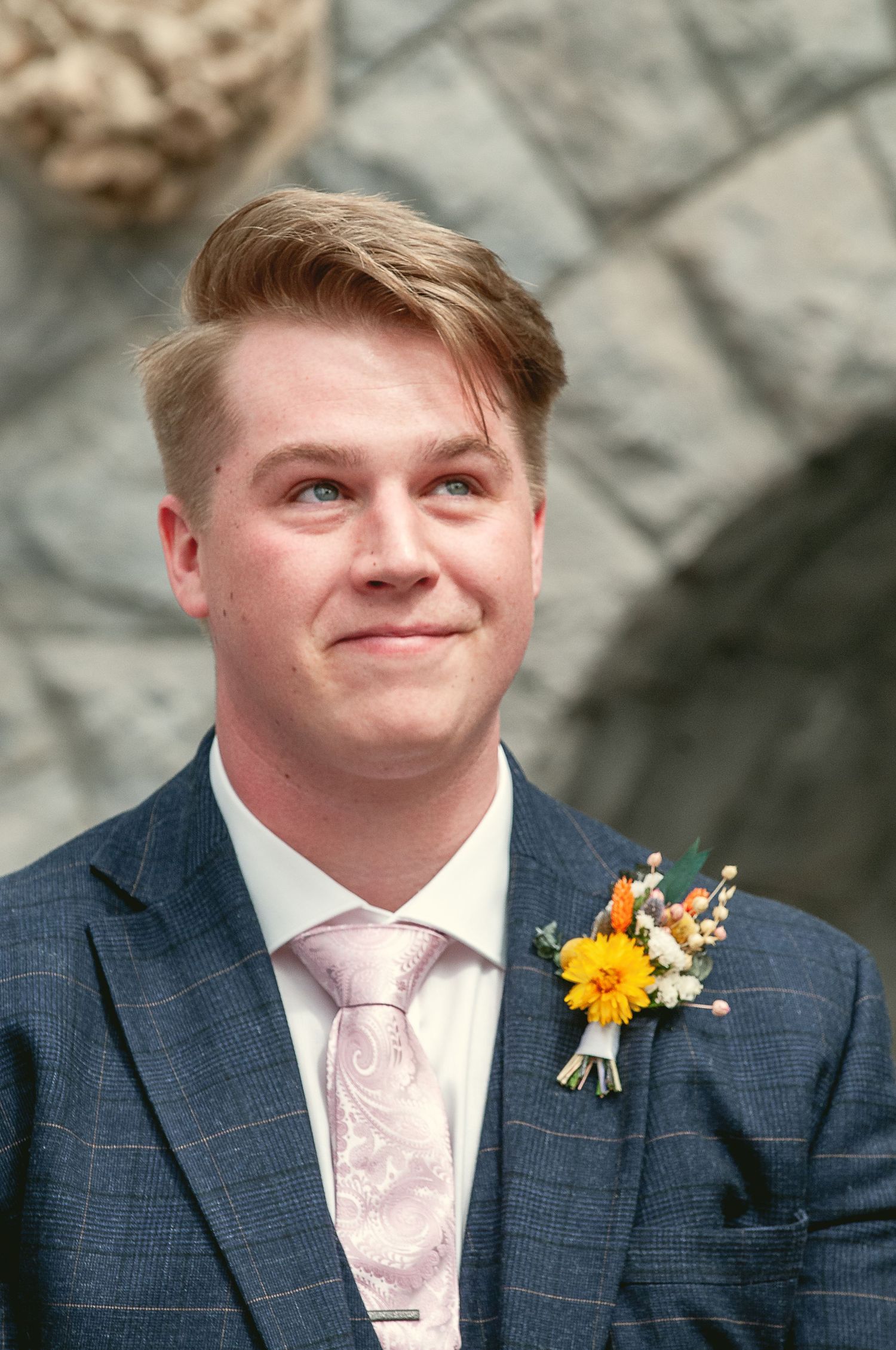 Groom wearing navy suit with pink tie and yellow boutonniere against stone wall.