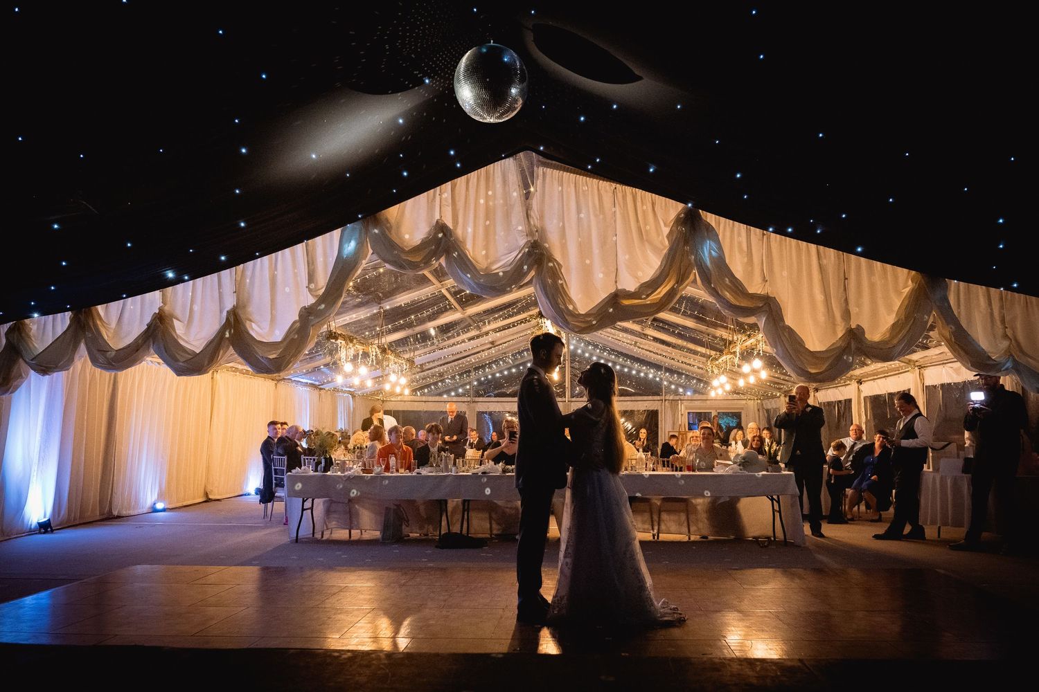 Couple shares first dance under starlit tent ceiling with elegant draped decor and glowing chandeliers at wedding reception.