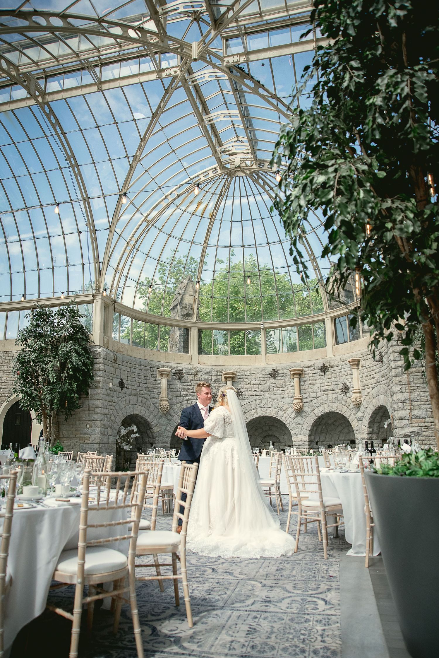 A romantic wedding scene in a stunning glass-domed conservatory with stone walls and elegant table settings.