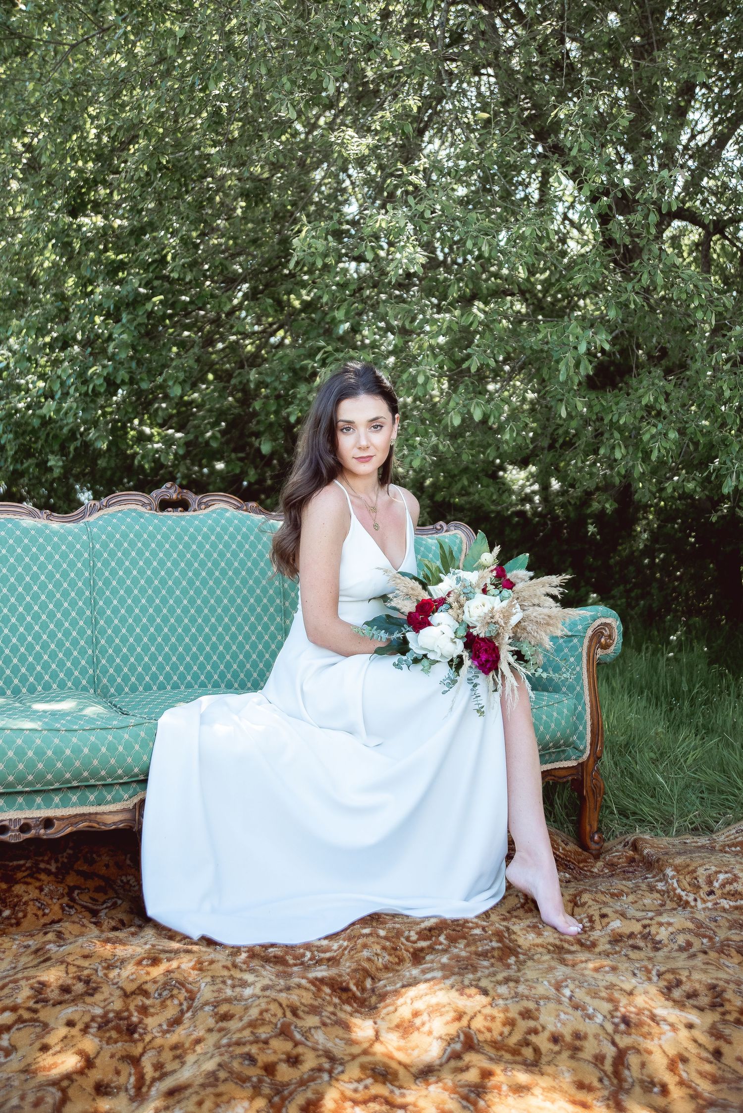A bride in white reclines on a mint green vintage sofa surrounded by fallen leaves in a garden setting.