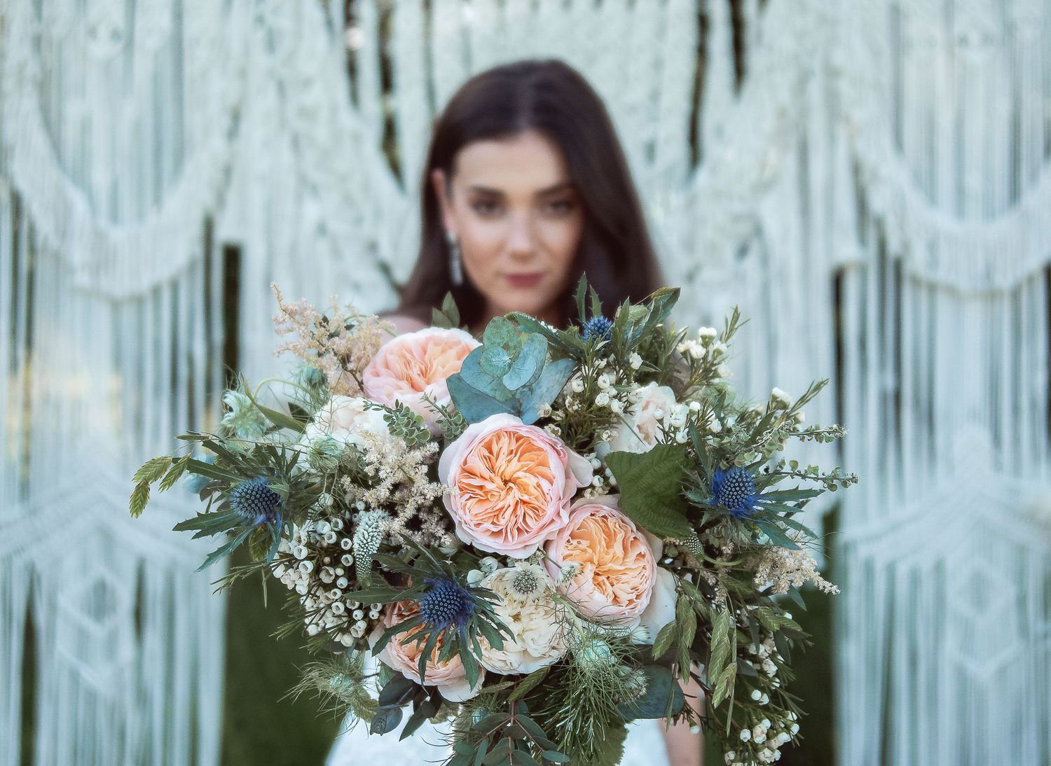 Close-up of a romantic peach and blue wedding bouquet with macrame backdrop.