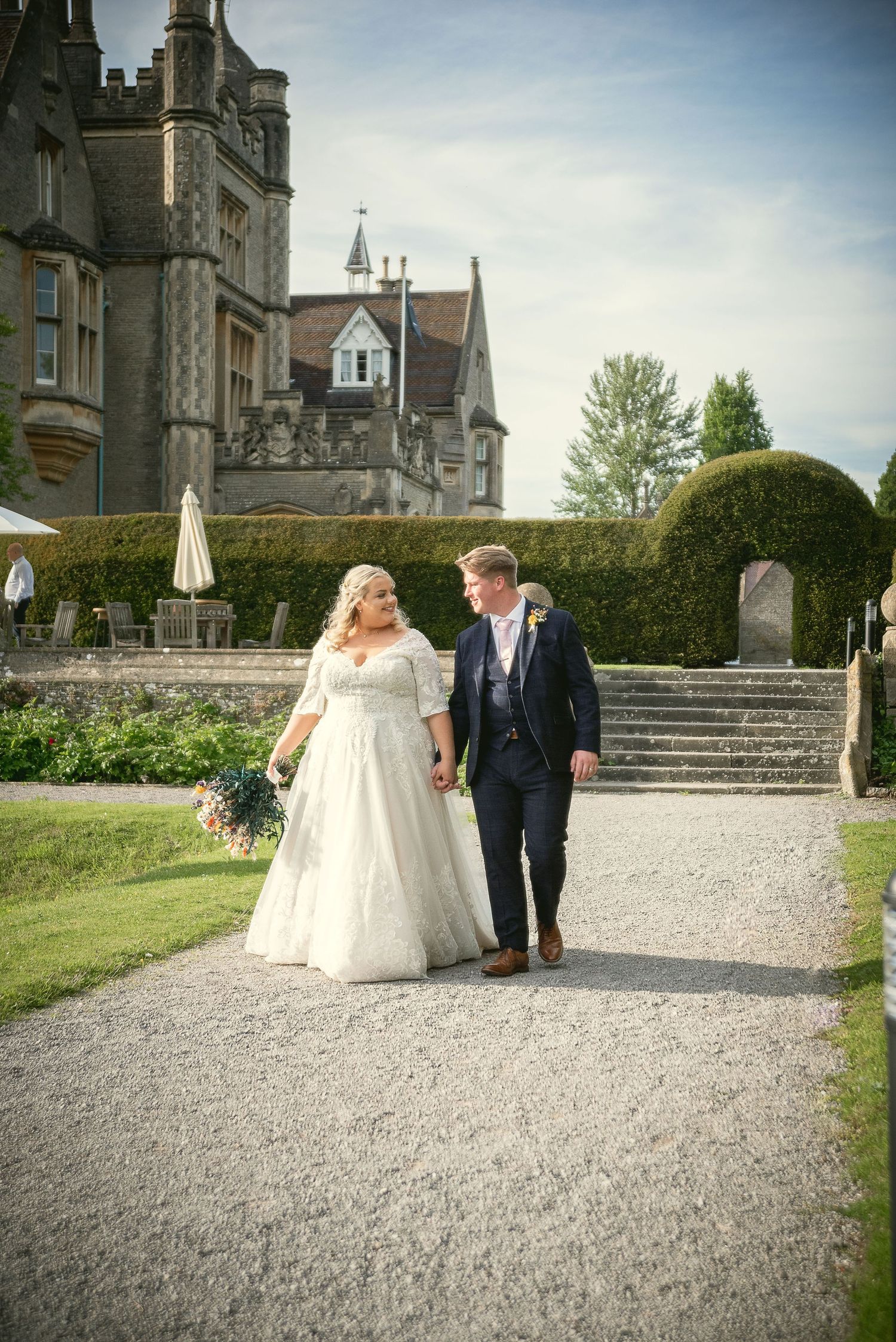 A wedding couple walks along a gravel path outside a grand stone castle on their special day.