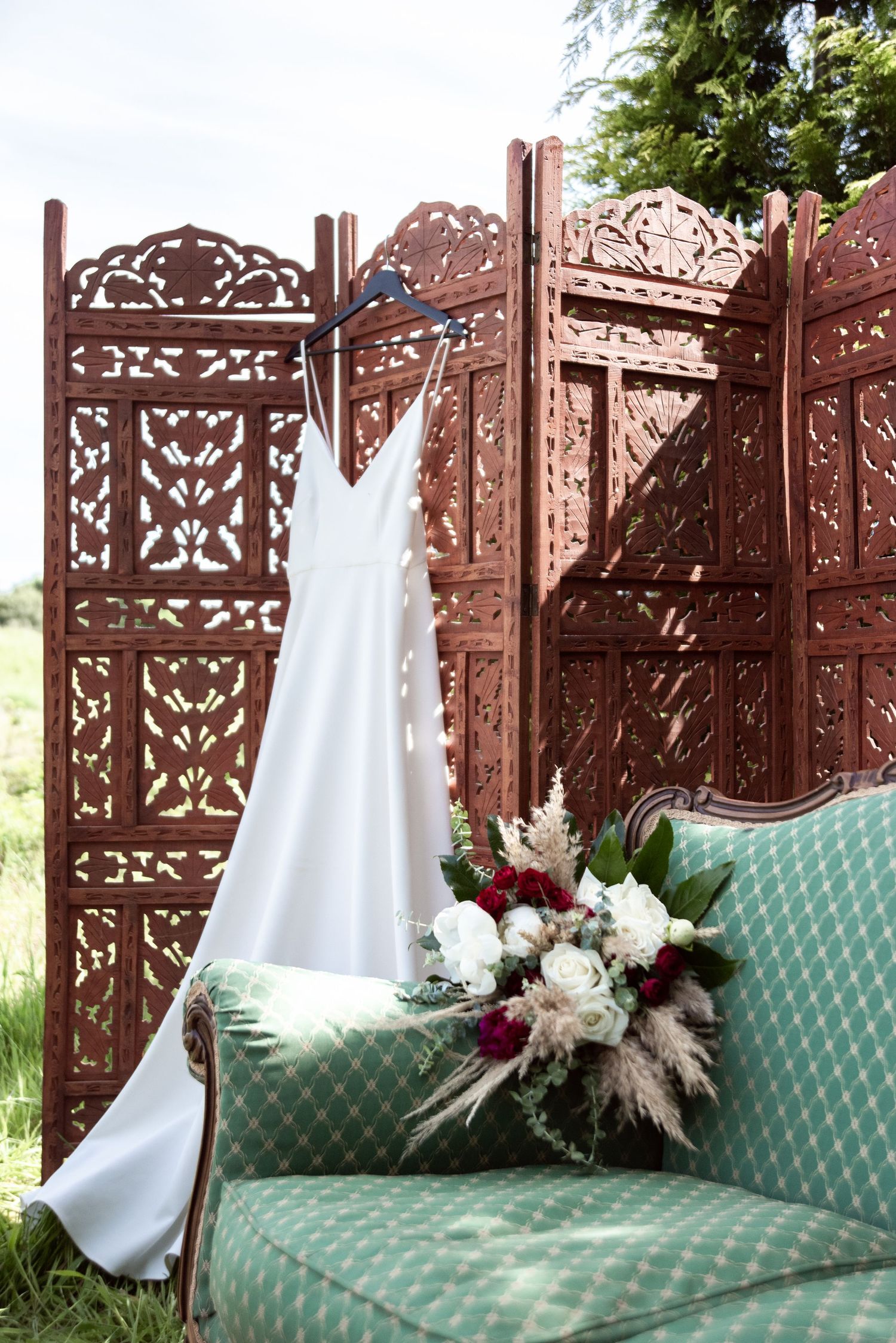 An elegant white dress and floral bouquet displayed on a mint green sofa against an ornate wooden screen backdrop.