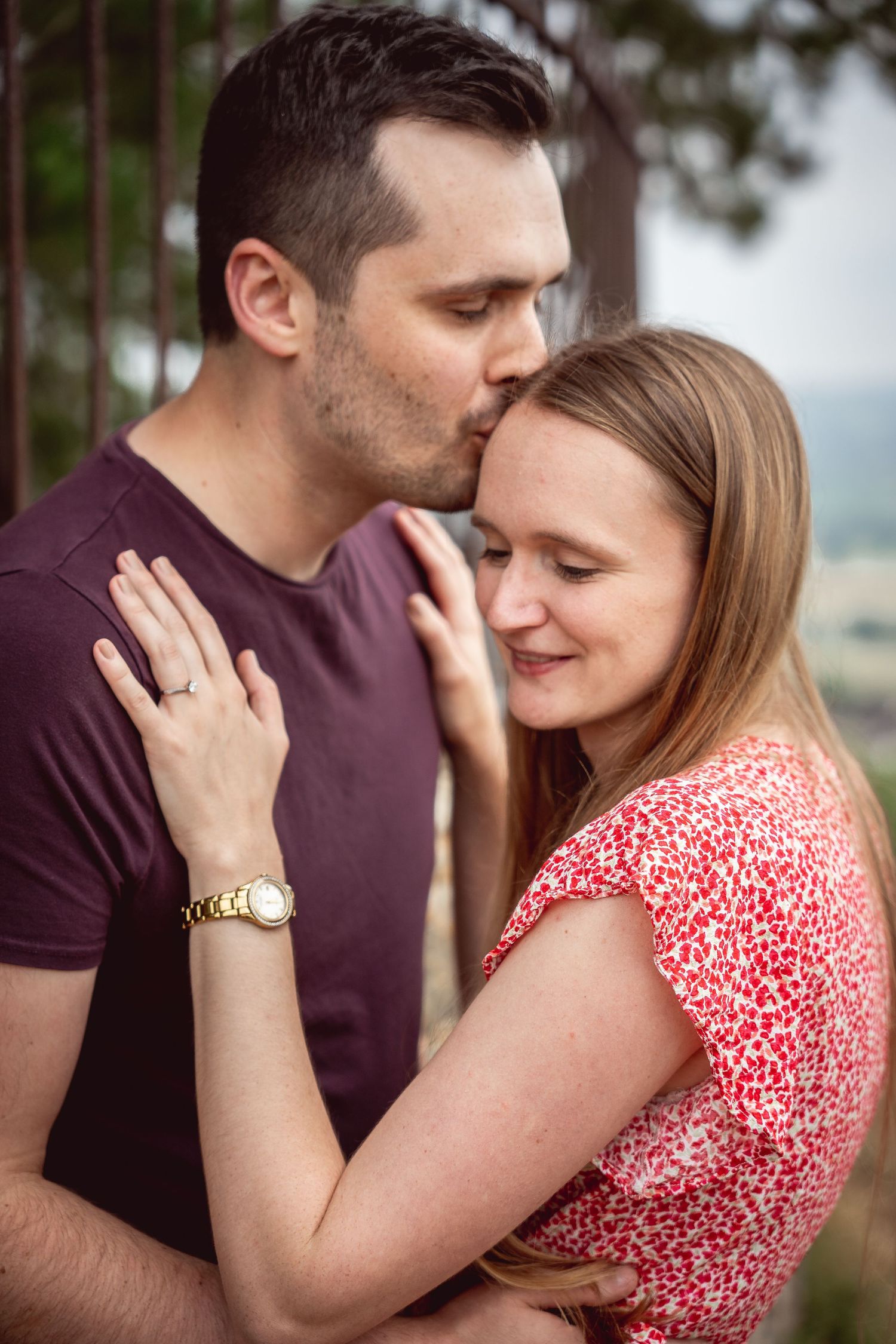man kissing woman on the head, woman with hands placed on his chest showing the engagement ring