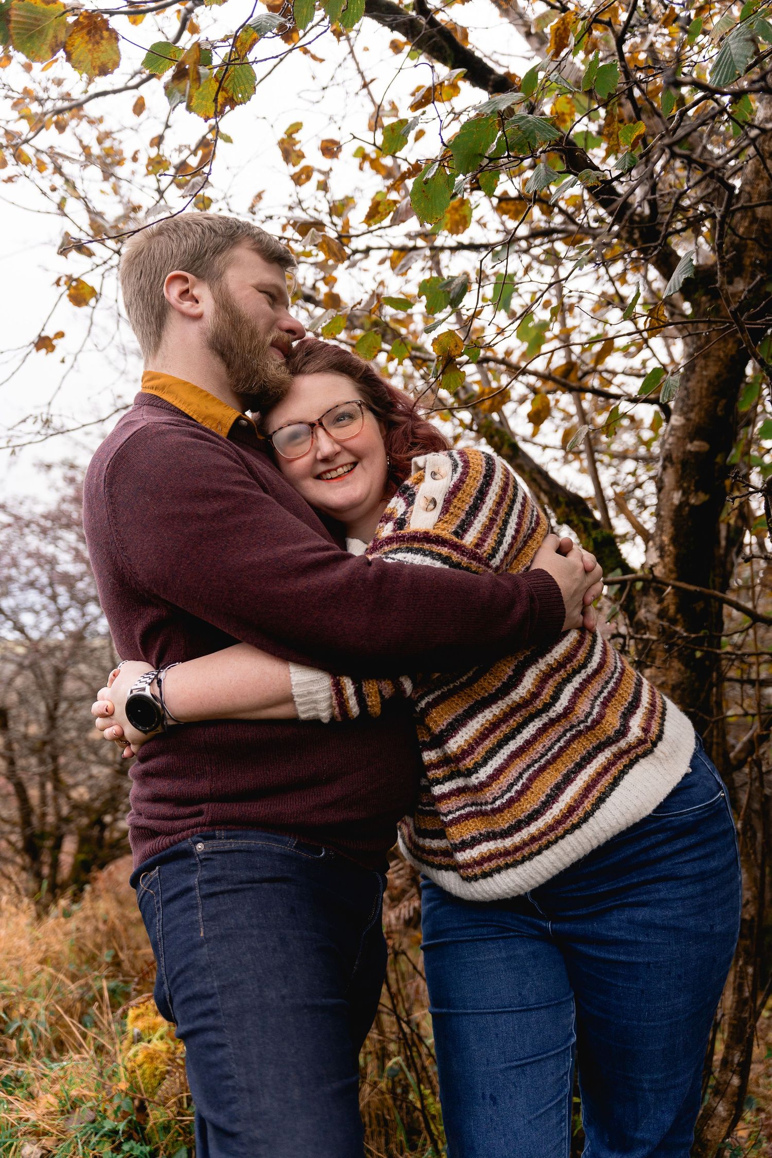 couple embracing under autumn trees as they take shelter from the rain, woman's glasses are fogging up  but she's smiling