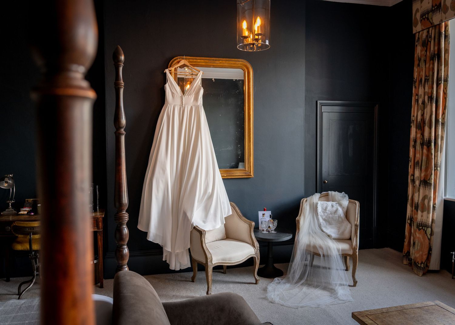 Wedding dress hangs elegantly in preparation room with natural light streaming through windows.