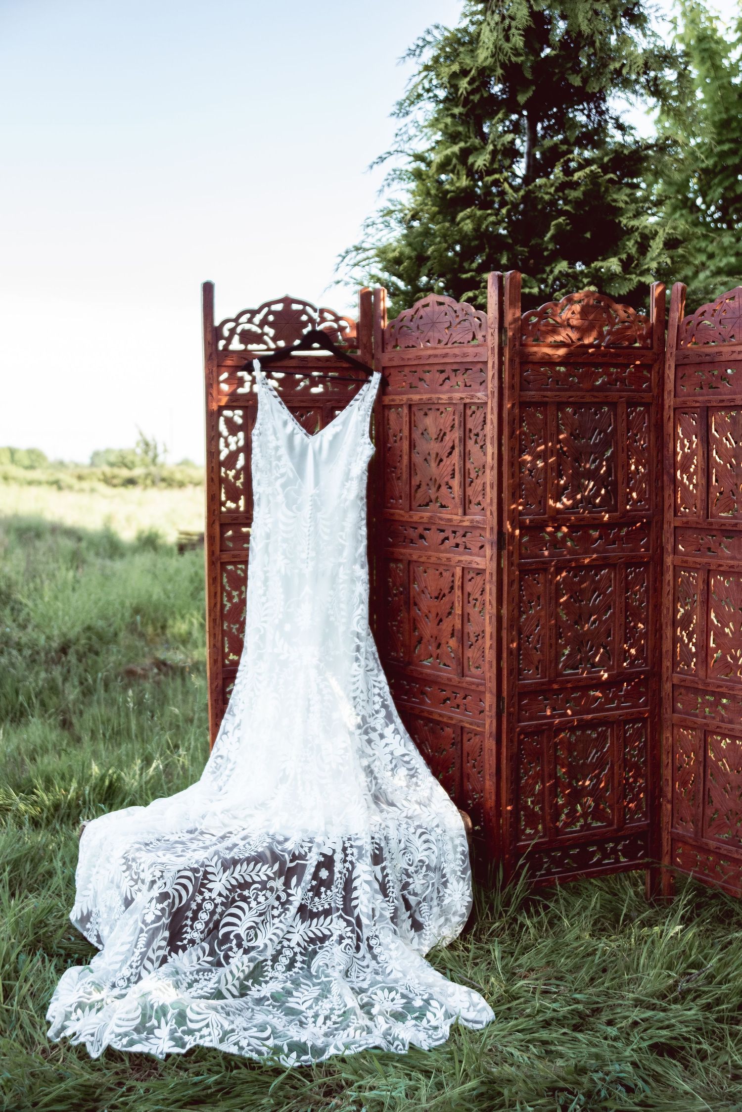 White lace wedding dress displayed against an ornate wooden screen backdrop outdoors.