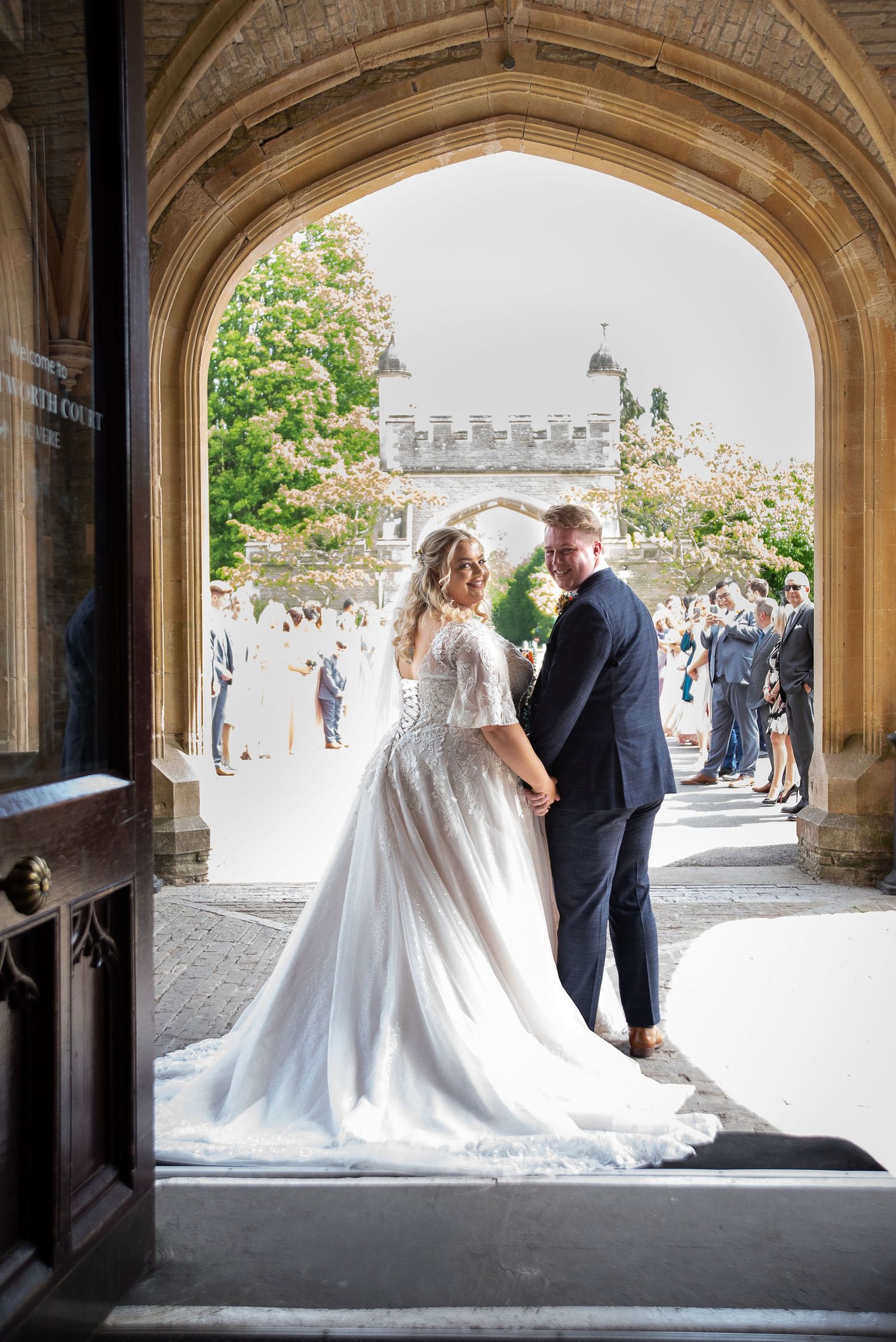 A wedding couple poses under a grand stone archway with guests visible in the background on a sunny day.