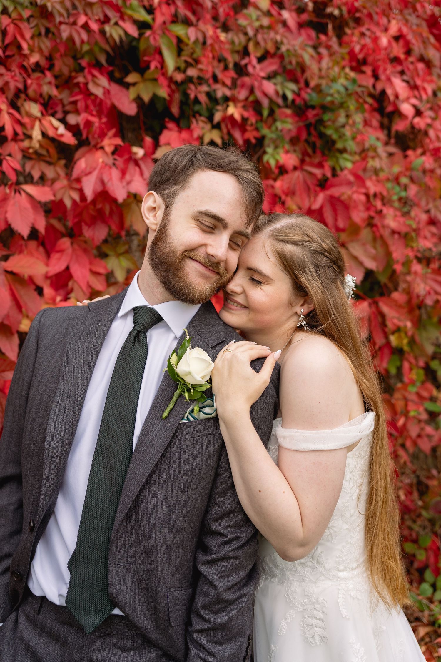Couple embracing in front of vibrant red autumn foliage, groom in gray suit, bride in white off-shoulder wedding dress.