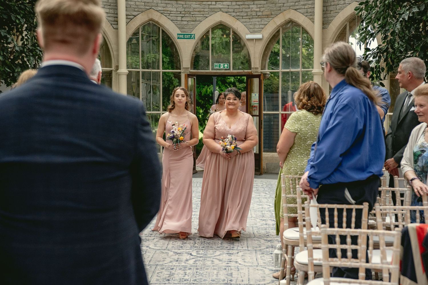 Bridesmaids in blush pink dresses standing at an outdoor wedding ceremony with gothic architecture.