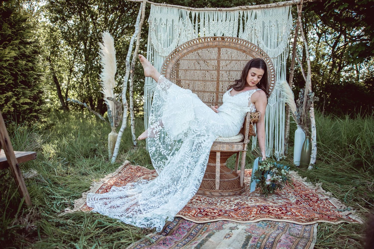 Outdoor bridal photoshoot setup with macrame backdrop, rugs, and vintage furniture in a garden setting.