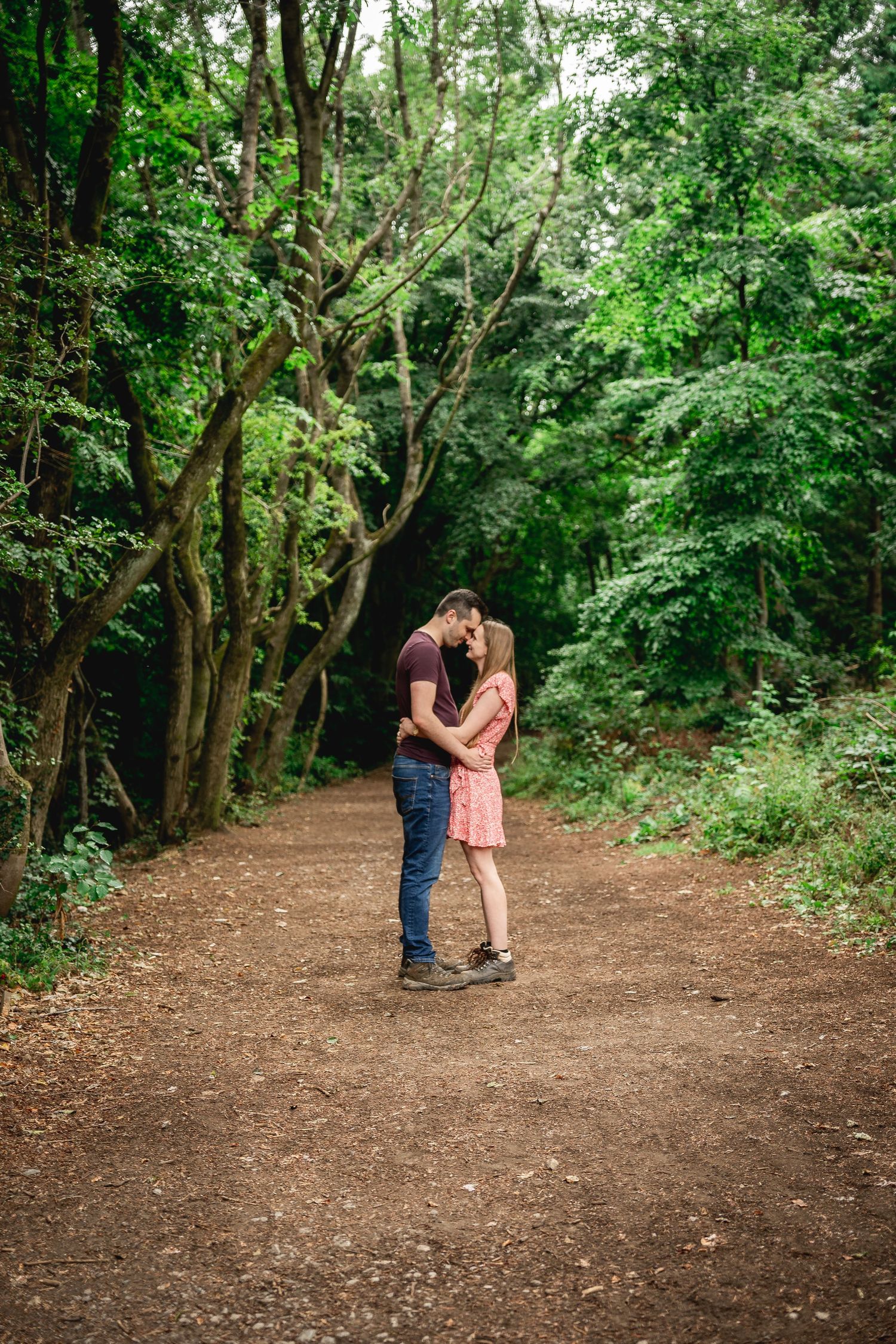 wide shot of couple standing facing each other with their foreheads touching in the middle of a woodland path
