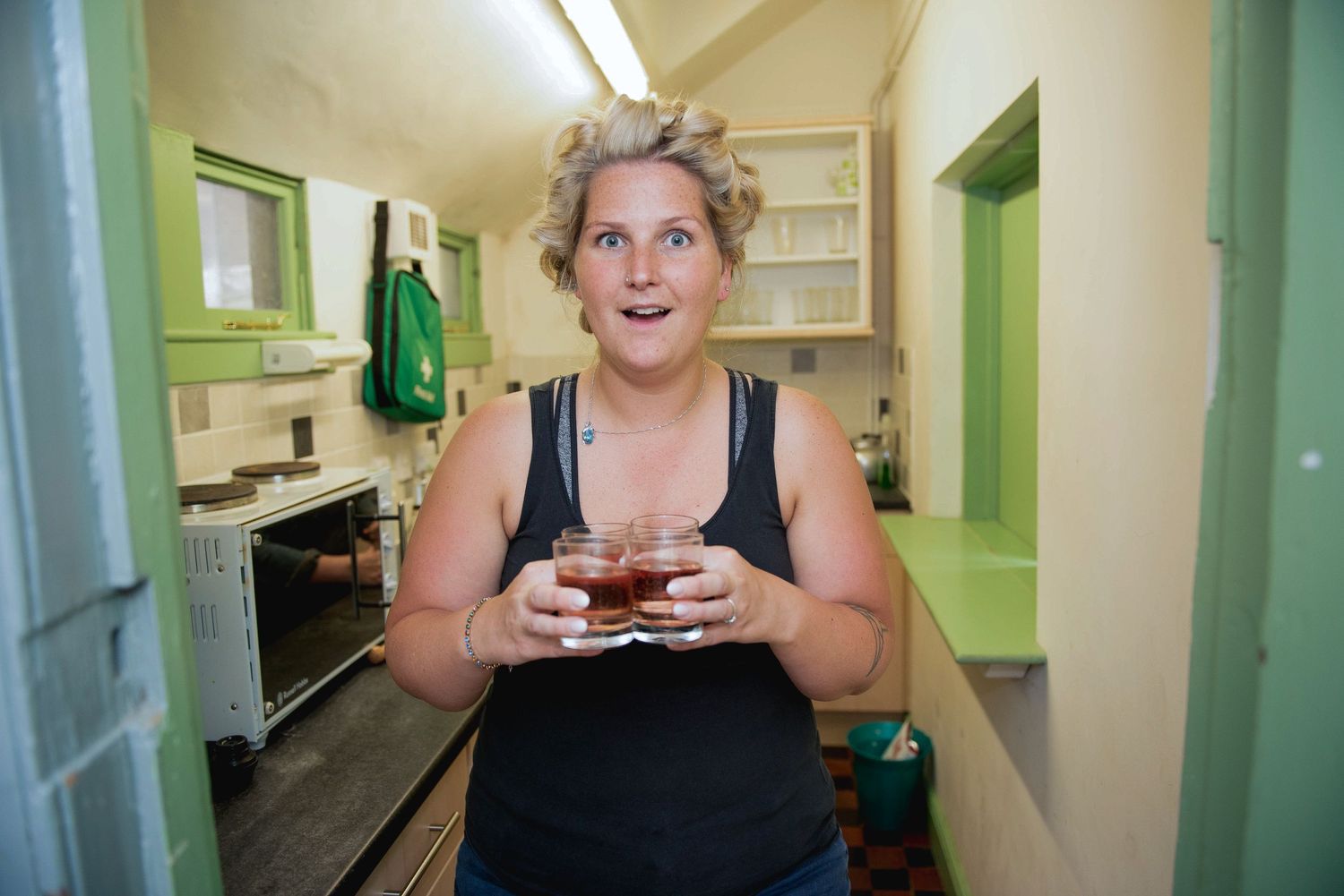 Bride with rollers in her hair holding large shot glasses looking surprised leaving the kitchen