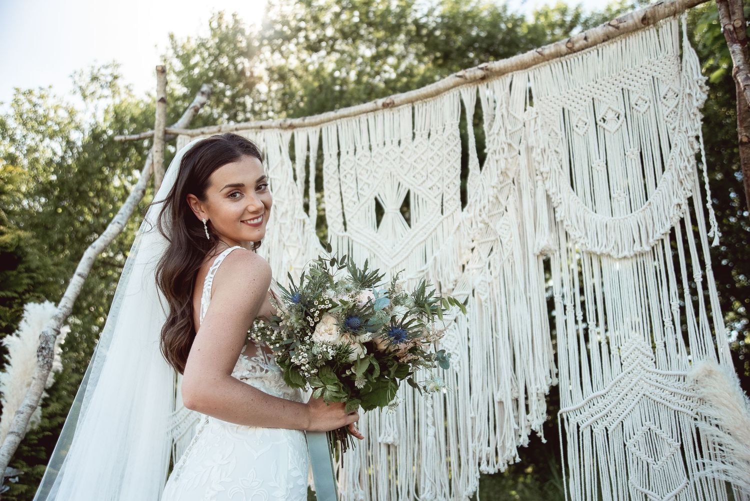 A bride in a lace wedding dress poses with a bouquet against a macrame backdrop in an outdoor garden setting.