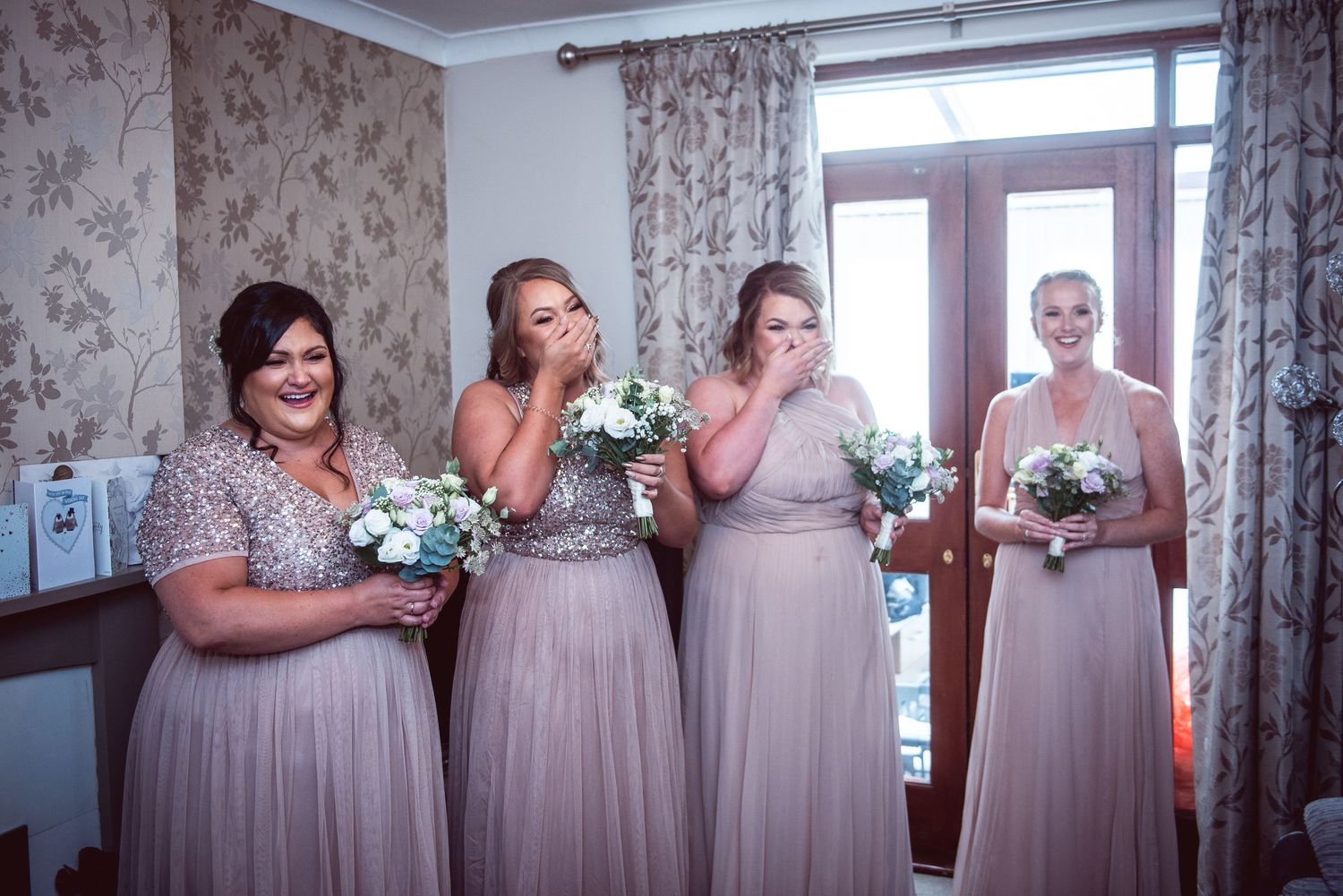 Lined up bridesmaids in pink dresses and holding bouquets reacting as the bride reveals her dress