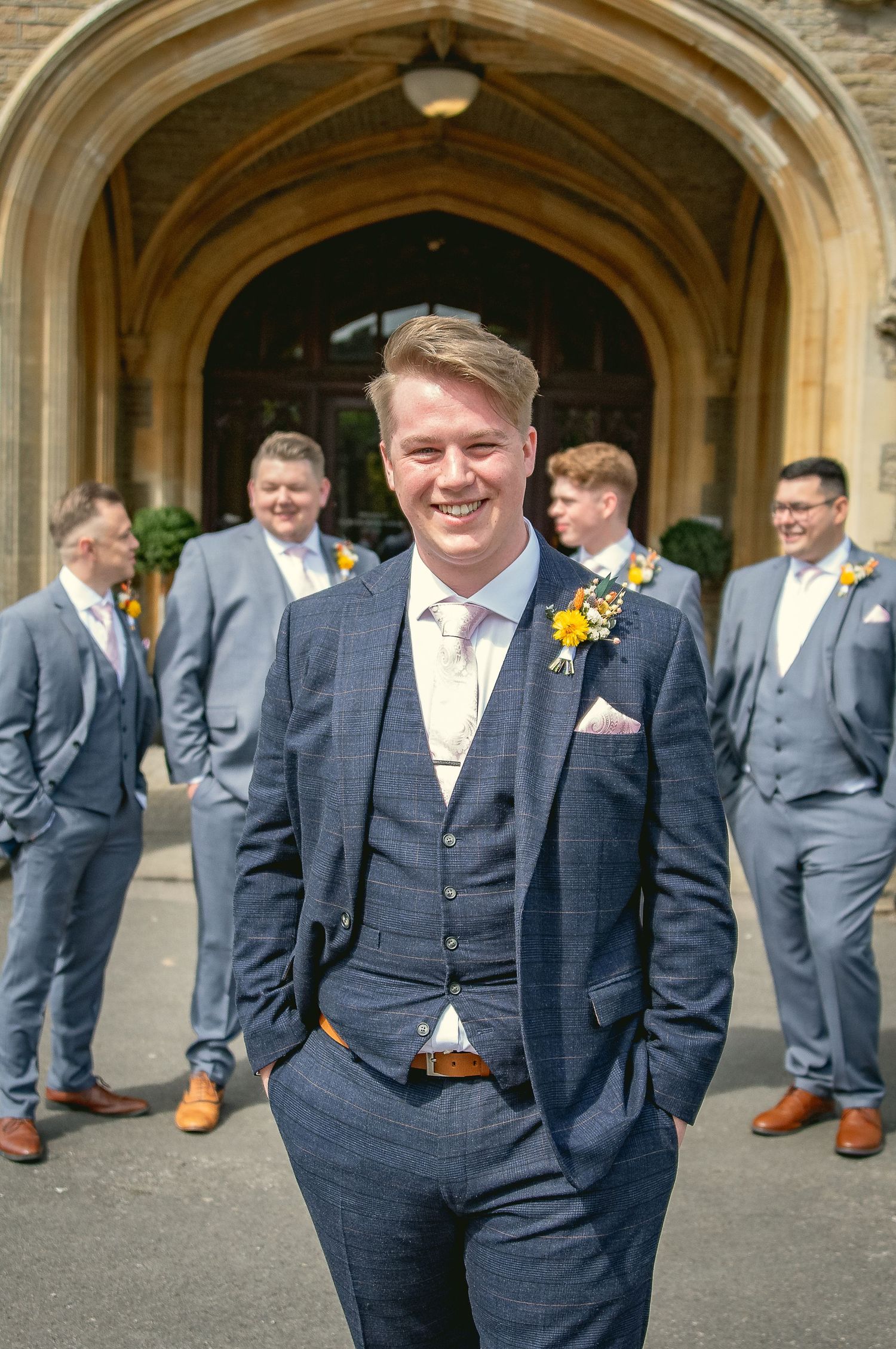 Groomsmen in navy suits and yellow ties gather under stone archways at an elegant wedding venue.