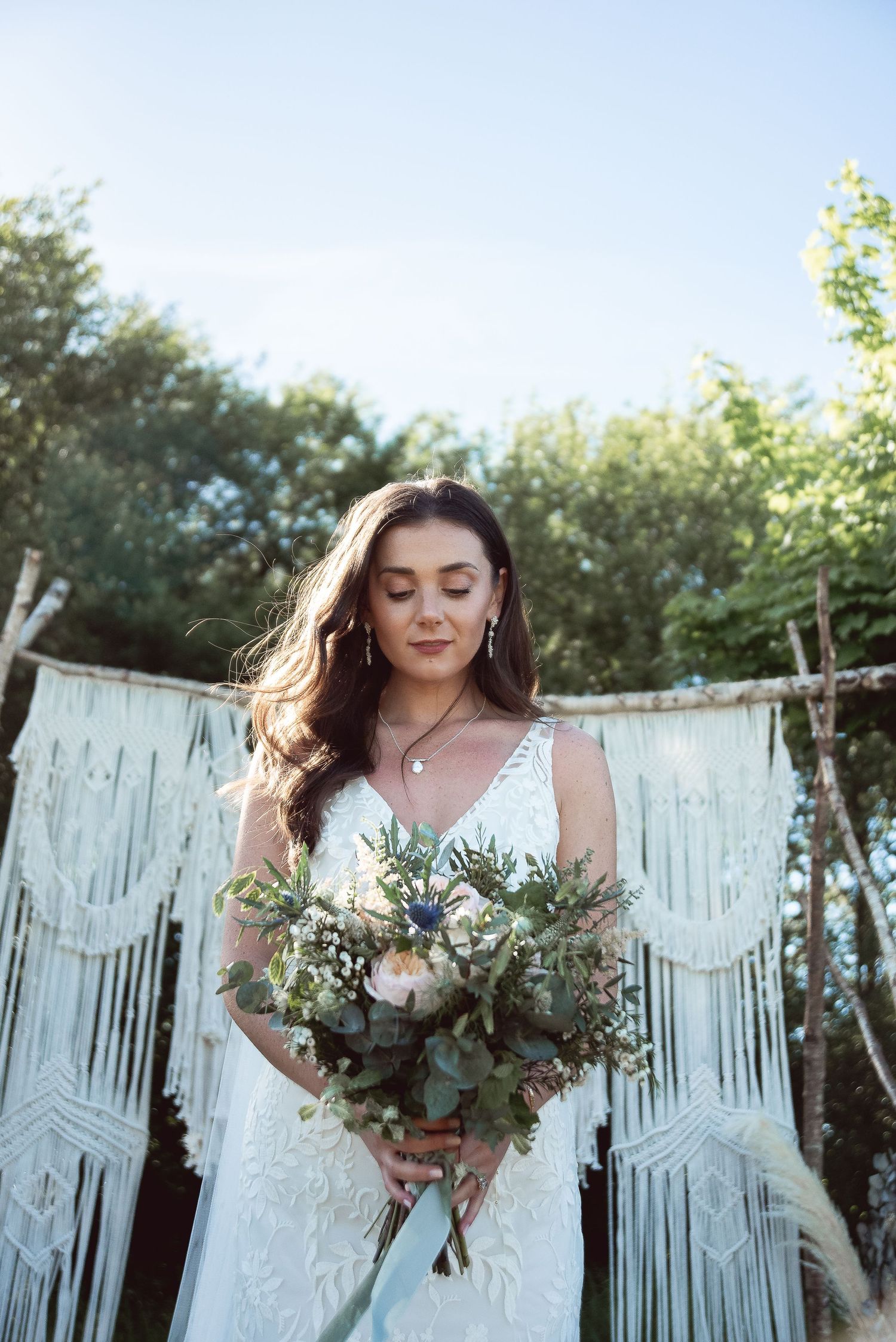 Outdoor bridal portrait with a flowing white dress and rustic macrame backdrop in a garden setting.